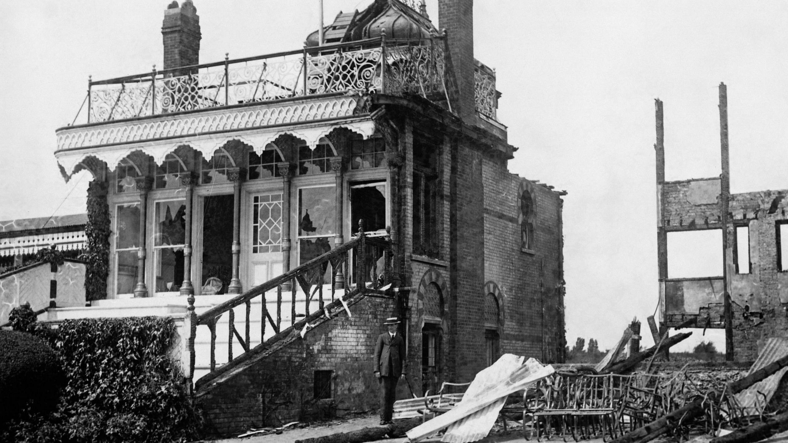 The remains of the Royal Box at Hurst Park grandstand in Molesey after it was burnt down in a protest by suffragettes. A man in a suit and a straw boater stands in front of the badly damaged building.