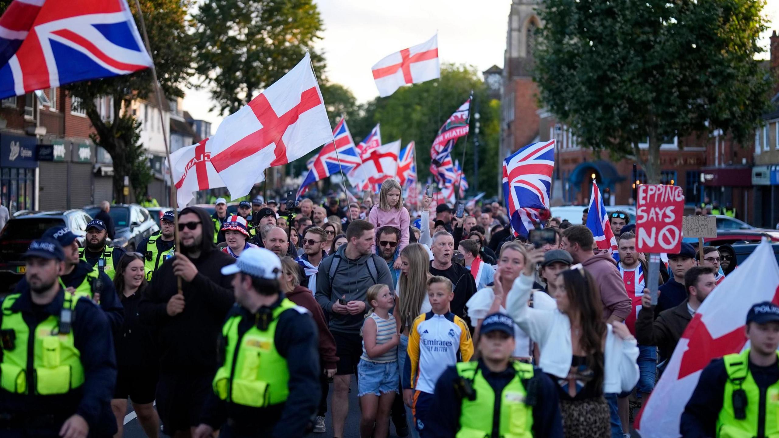 Protesters marching in a tight group, marshalled by a line of police officers at the front. The protesters are waving England and Union flags. There are a handful of women and children near the front of the crowd.