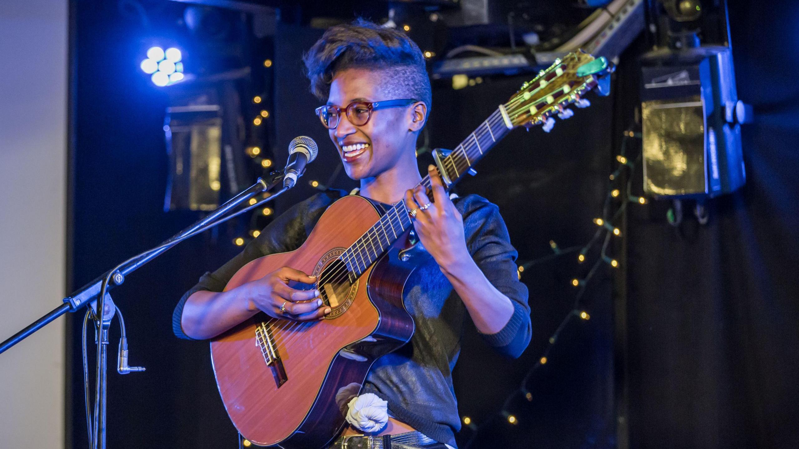 A musician stands on a small stage holding an acoustic guitar beside a microphone, illuminated by blue stage lighting. The dark venue background is decorated with warm string lights, creating an intimate live-performance setting.