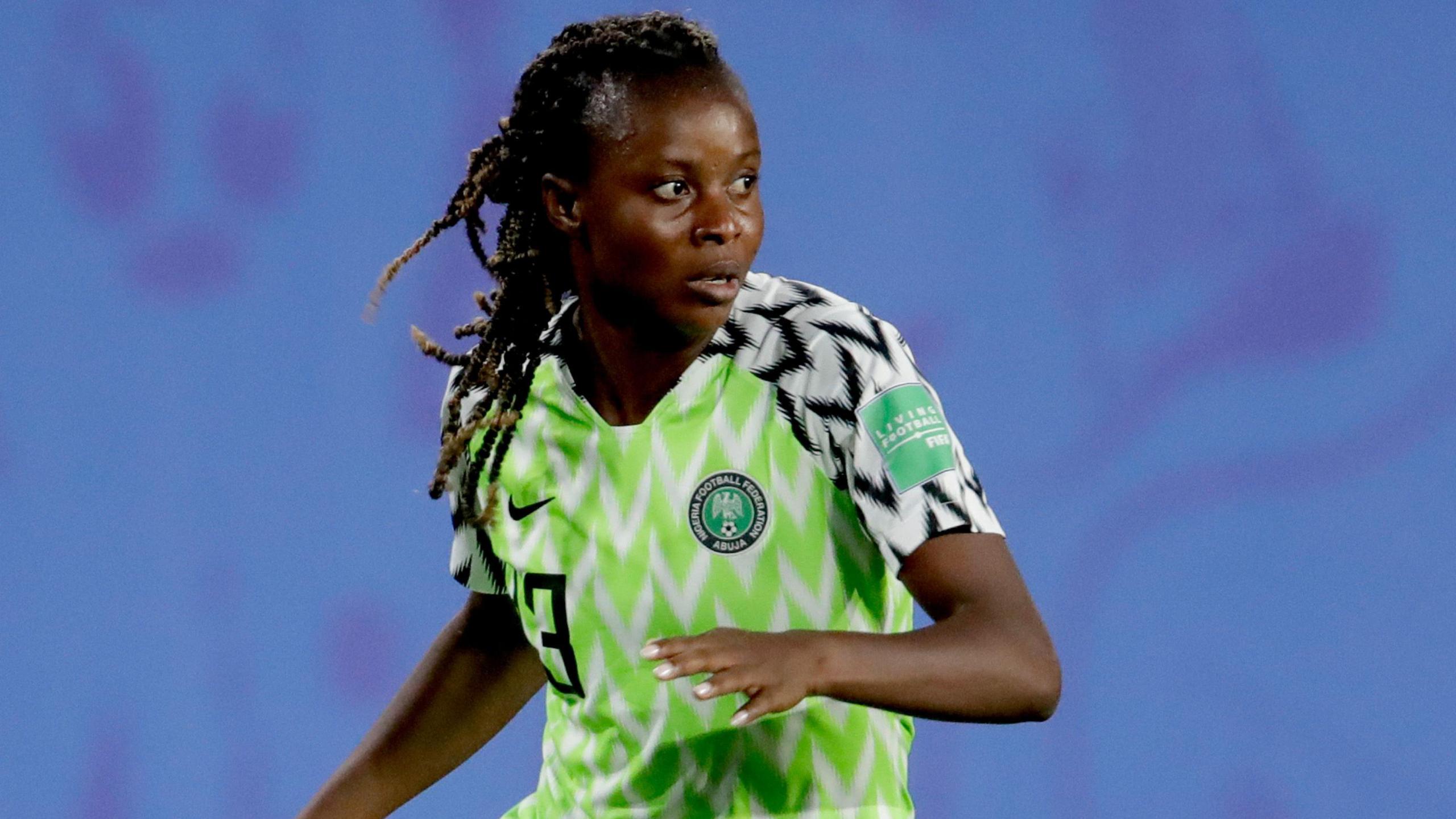 Ngozi Okobi-Okeoghene, visible from waist up wearing a green and white Nigeria shirt with black and white trim on the upper arms, raises her left arm to chest height and looks to her left during a football match