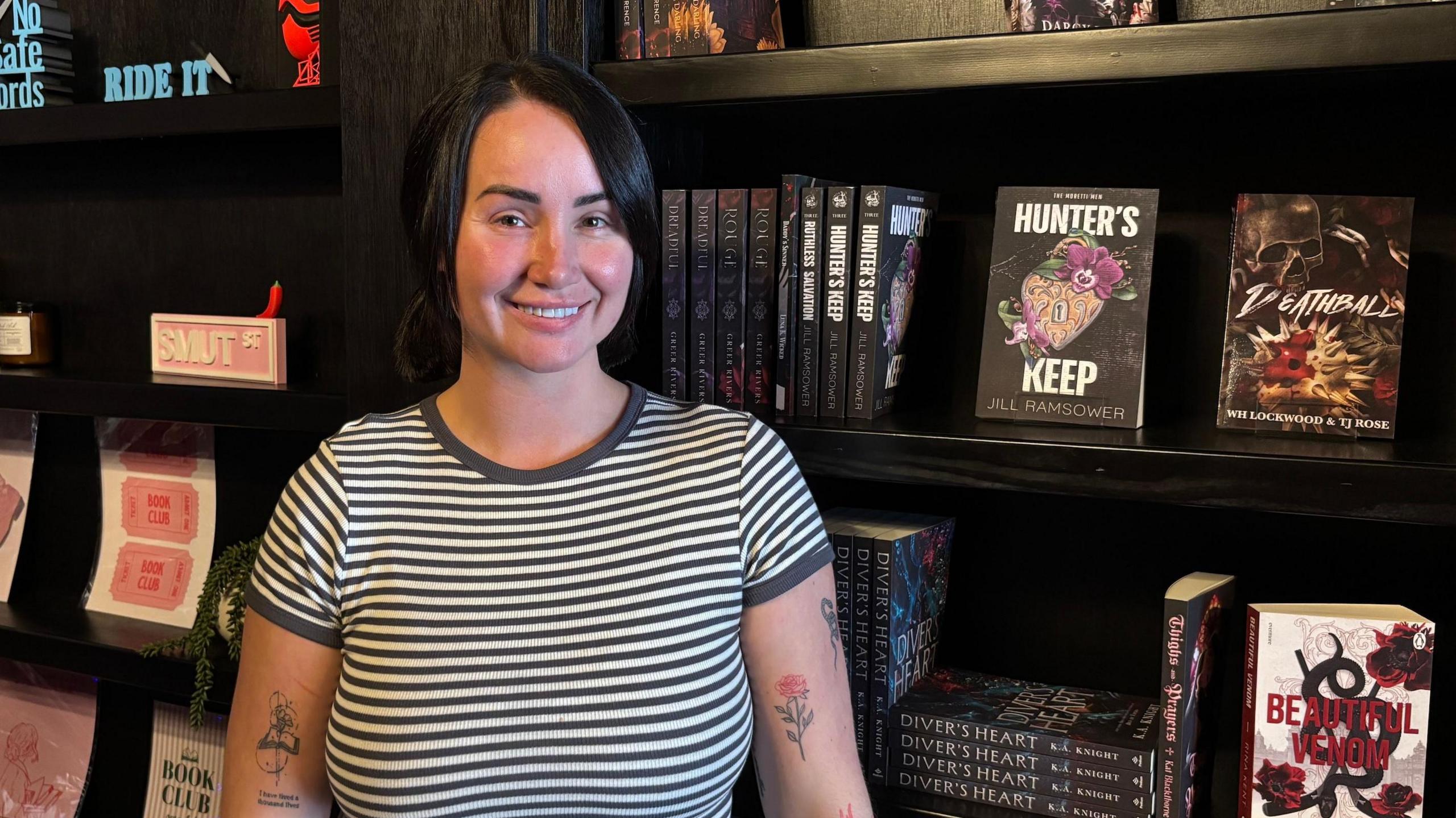 Ezibela Welsh, wearing a grey and white striped top. She has dark black hair that is tied into two pigtails at the back of her head, and side bangs. She has a small tattoo of a book on one arm, and two other small ones of a rose and a snake on the other. She is standing in front of her bookcases, which have an array of books from different sub-genres of romance.