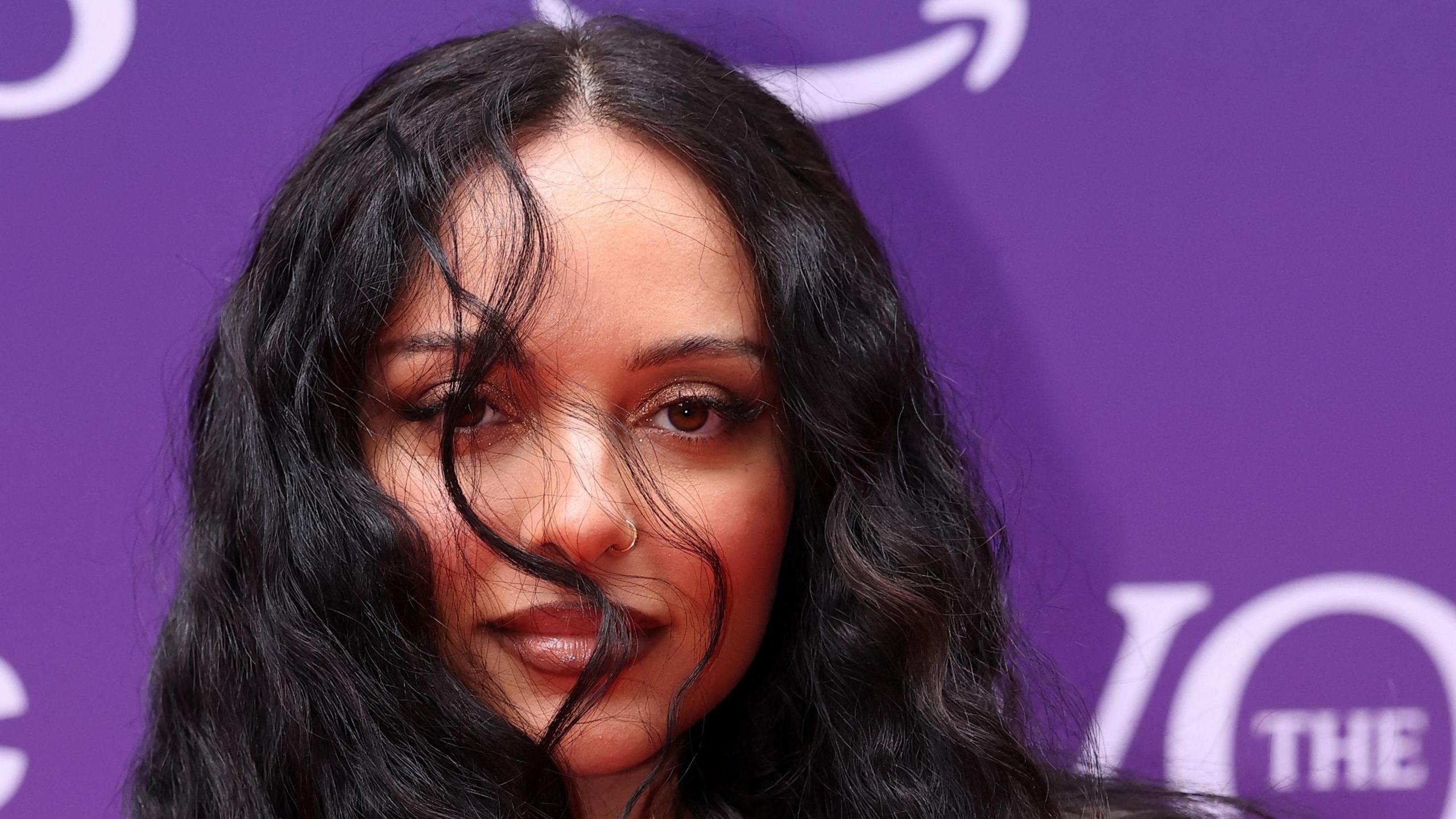 Jade Thirlwall is standing in front of a purple background with a few strands of her hair in front of her right eye.