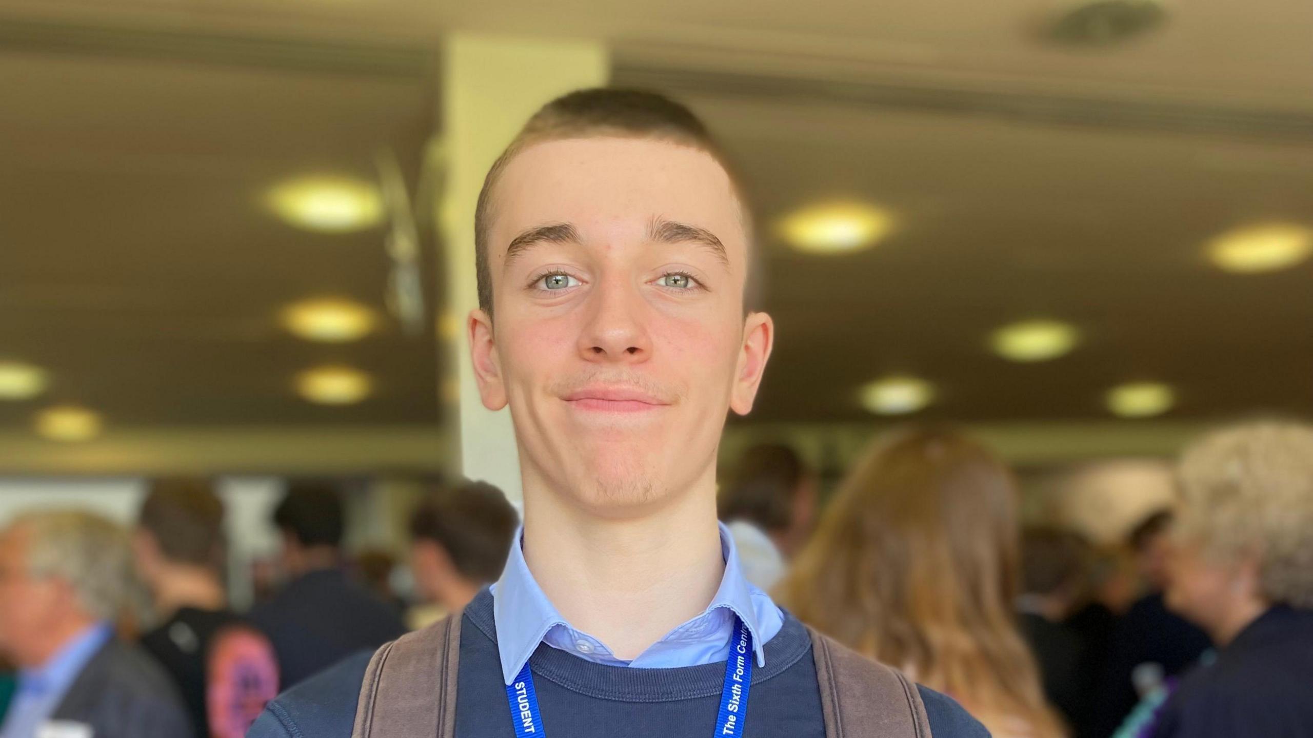A boy with short hair smiling towards the camera, he's wearing a brown backpack and you can see the straps on his shoulders. He's also wearing a lanyard and a blue jumper.