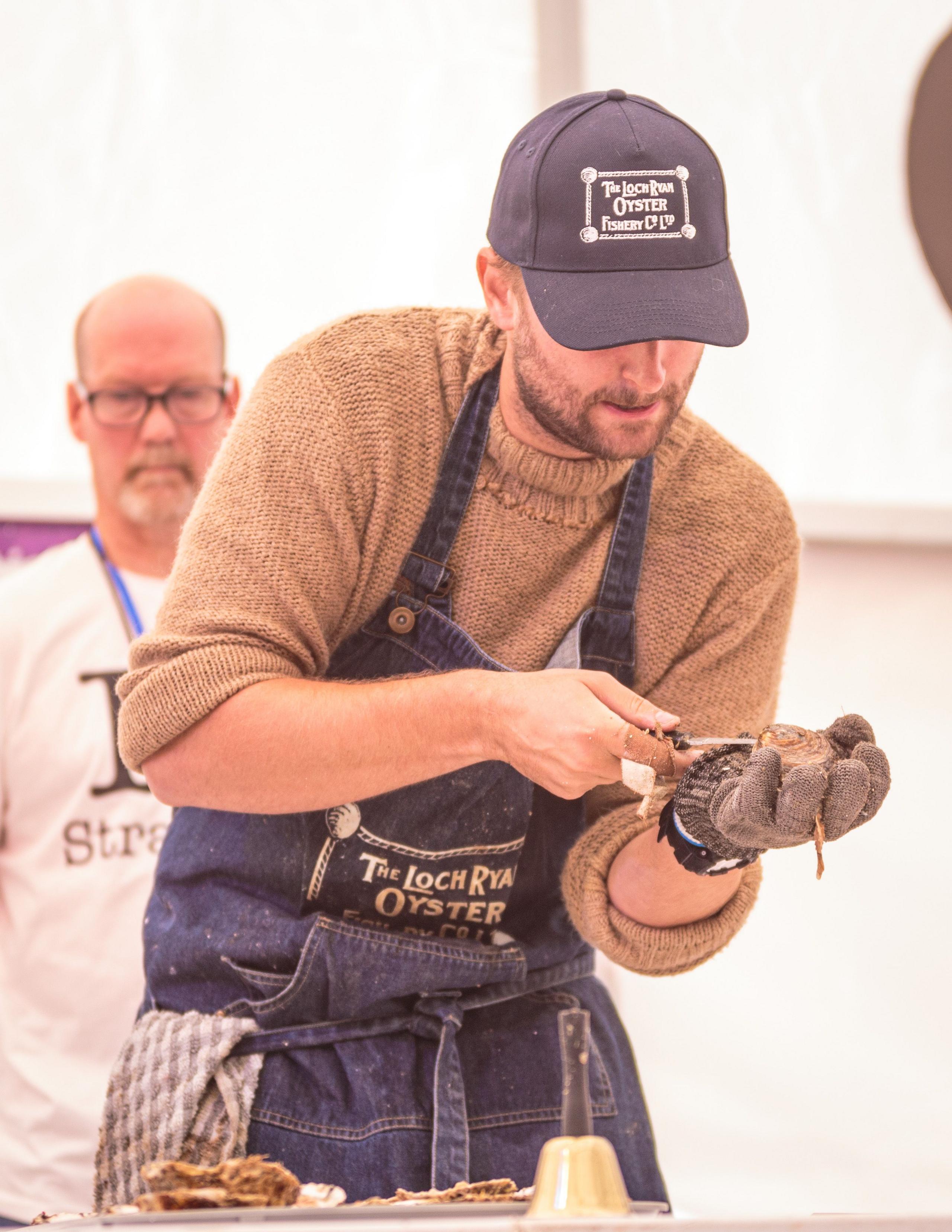 A man in a baseball cap and blue apron expertly shucks an oyster