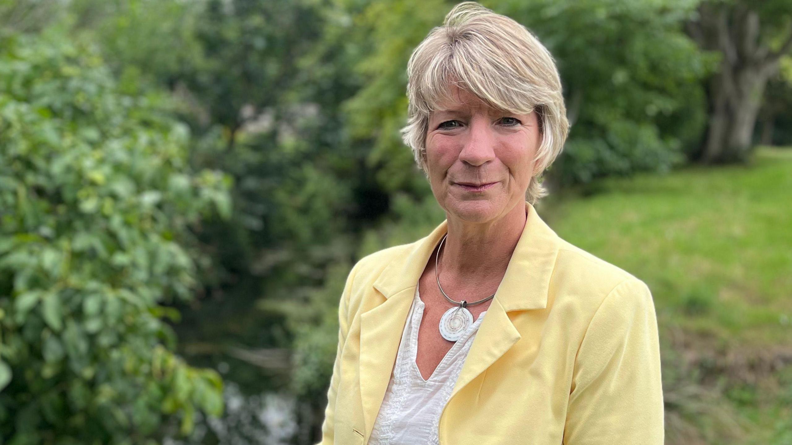 Pippa Heylings looks directly at the camera, smiling slightly. She is standing on a riverbank with a body of water behind her. She is wearing a pale yellow jacket and white top, with a silver necklace. She has short blonde hair.