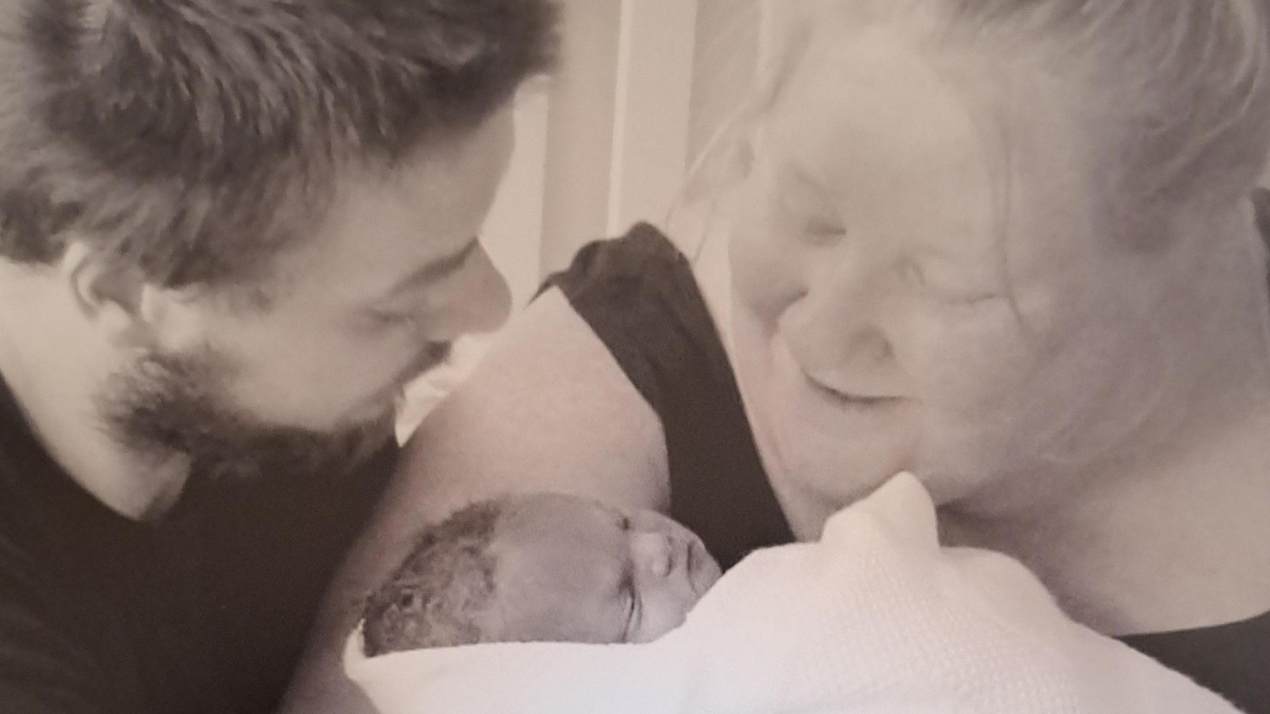 A black and white image of Lee and Kristina Pepper holding their son Theo who is wrapped in a swaddle