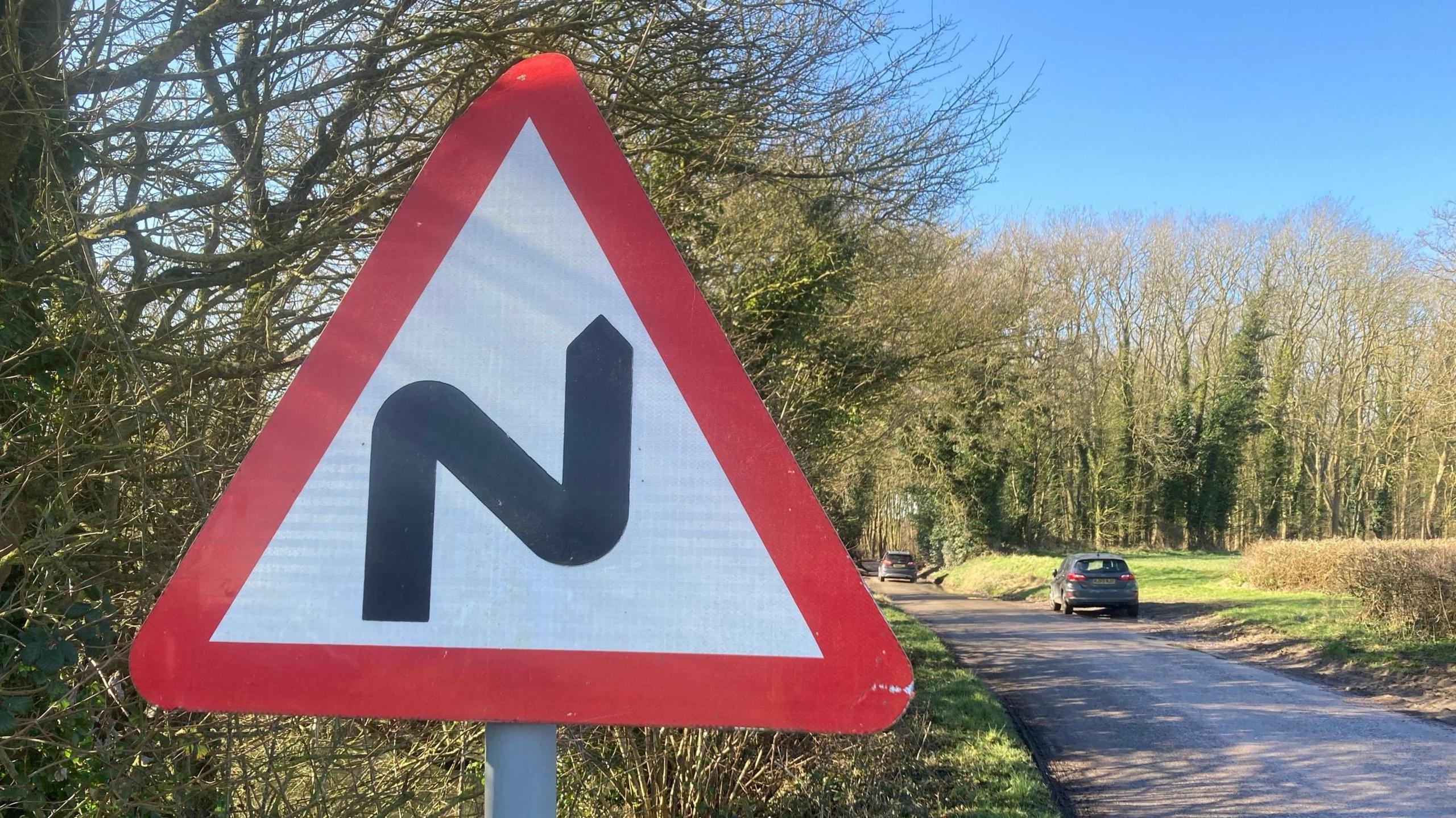 On the left of the picture is a triangular sign which has a red border, and white centre. It has a N shape on it which represents a bend in the road. On the right is he road leading towards the distance with 2 cars on it. 