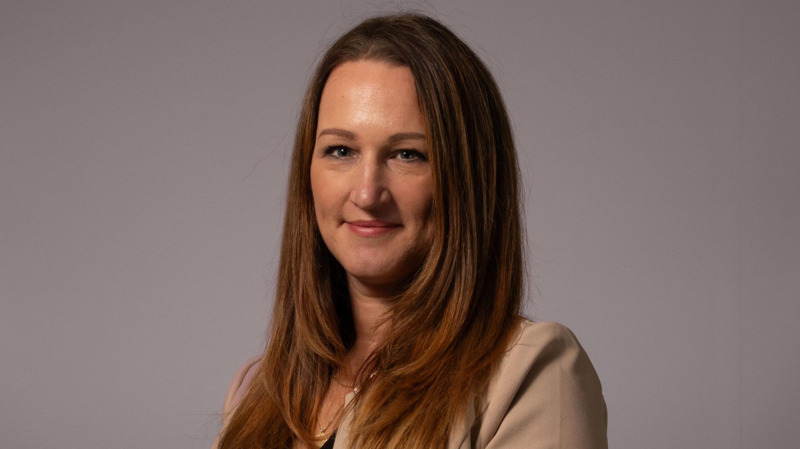 Kate, a smartly dressed woman wearing a beige suit, with long brown hair, smiles at the camera. It is a head and shoulders shot of her.