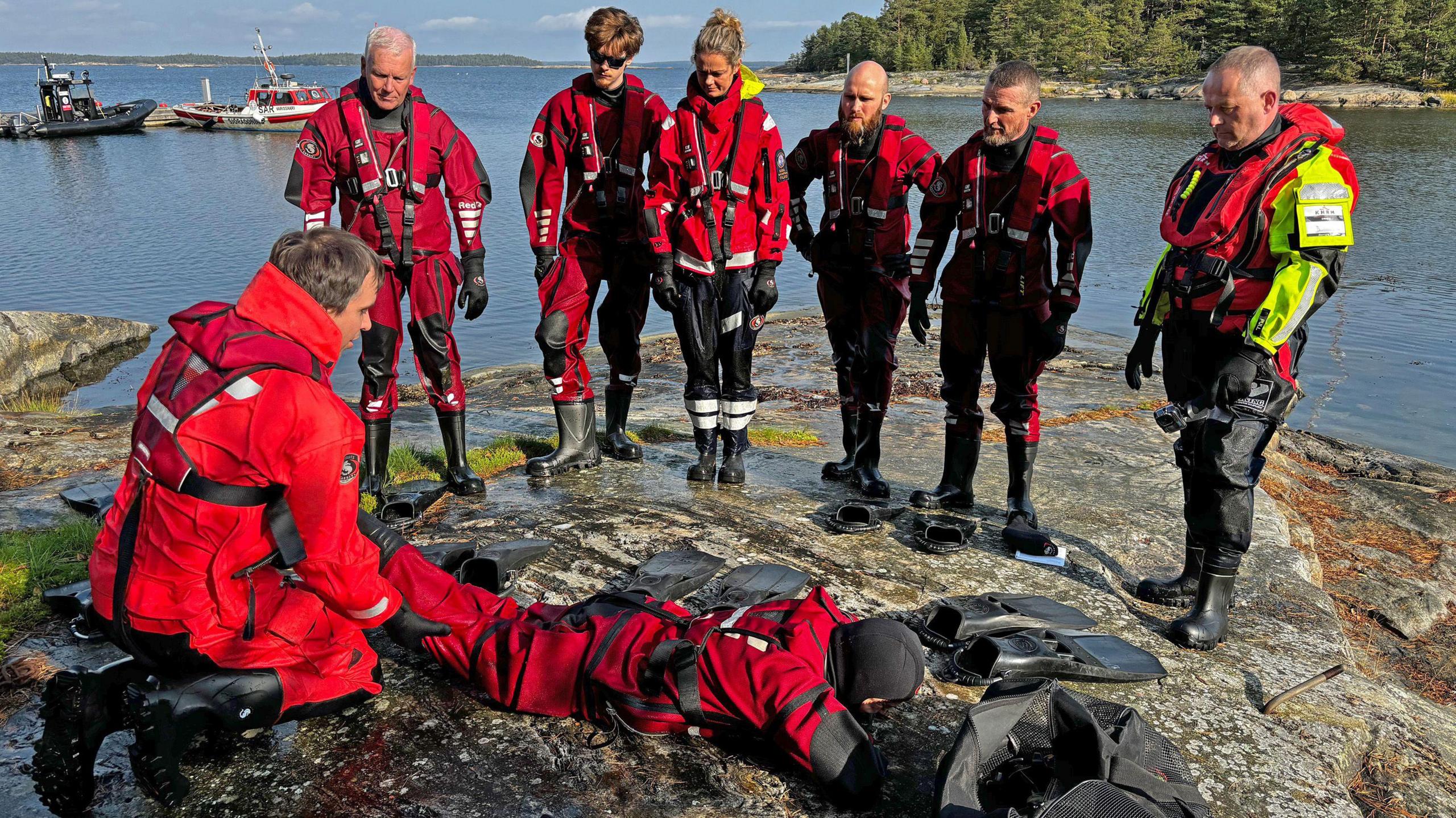 Six men and Tamara are standing on a rocky outcrop with the sea behind them. They are looking down at a man on his knees who is lifting the leg of another man who is lying down as he demonstrates a life-saving skill. All of them are wearing RNLI uniforms and buoyancy aids.