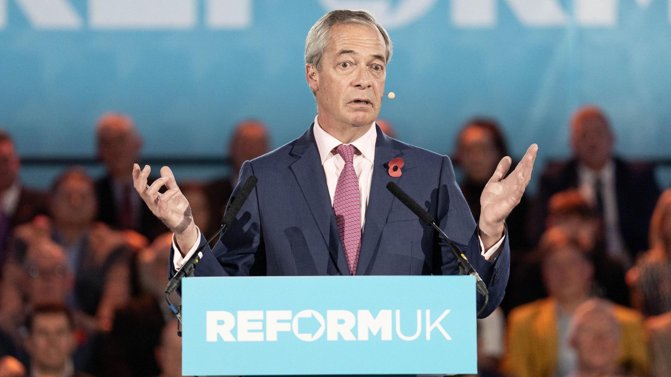 Nigel Farage in a suit and tie standing behind a podium with the words ‘REFORM UK’ printed on the front, speaking into two microphones. The background shows a large blue banner with partial text and an audience seated behind.