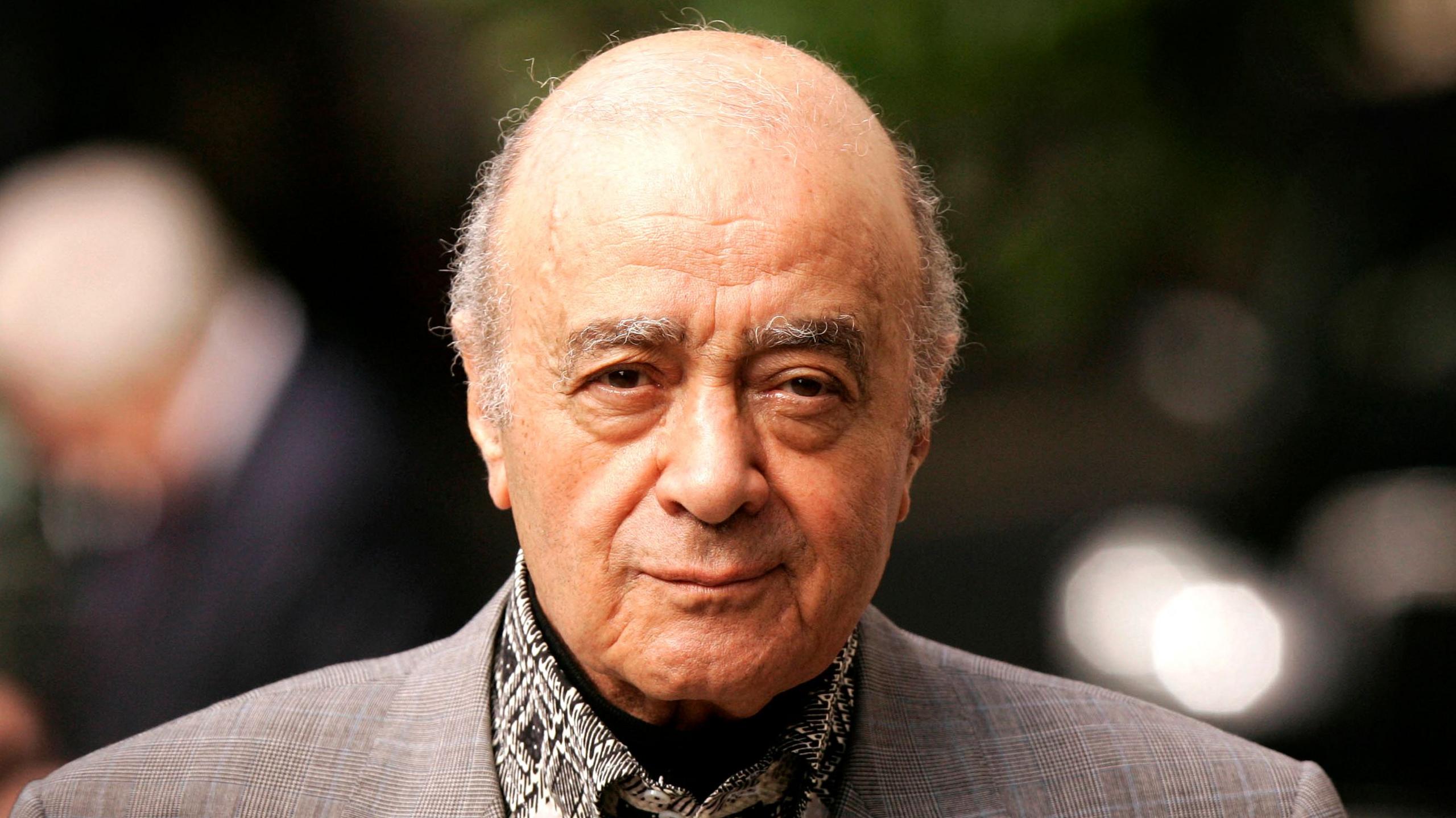 Headshot of Al Fayed, a balding man with a wrinkled face and greying eyebrows. He is looking at the camera wearing a grey suit jacket, black patterned shirt and a black cravat. 