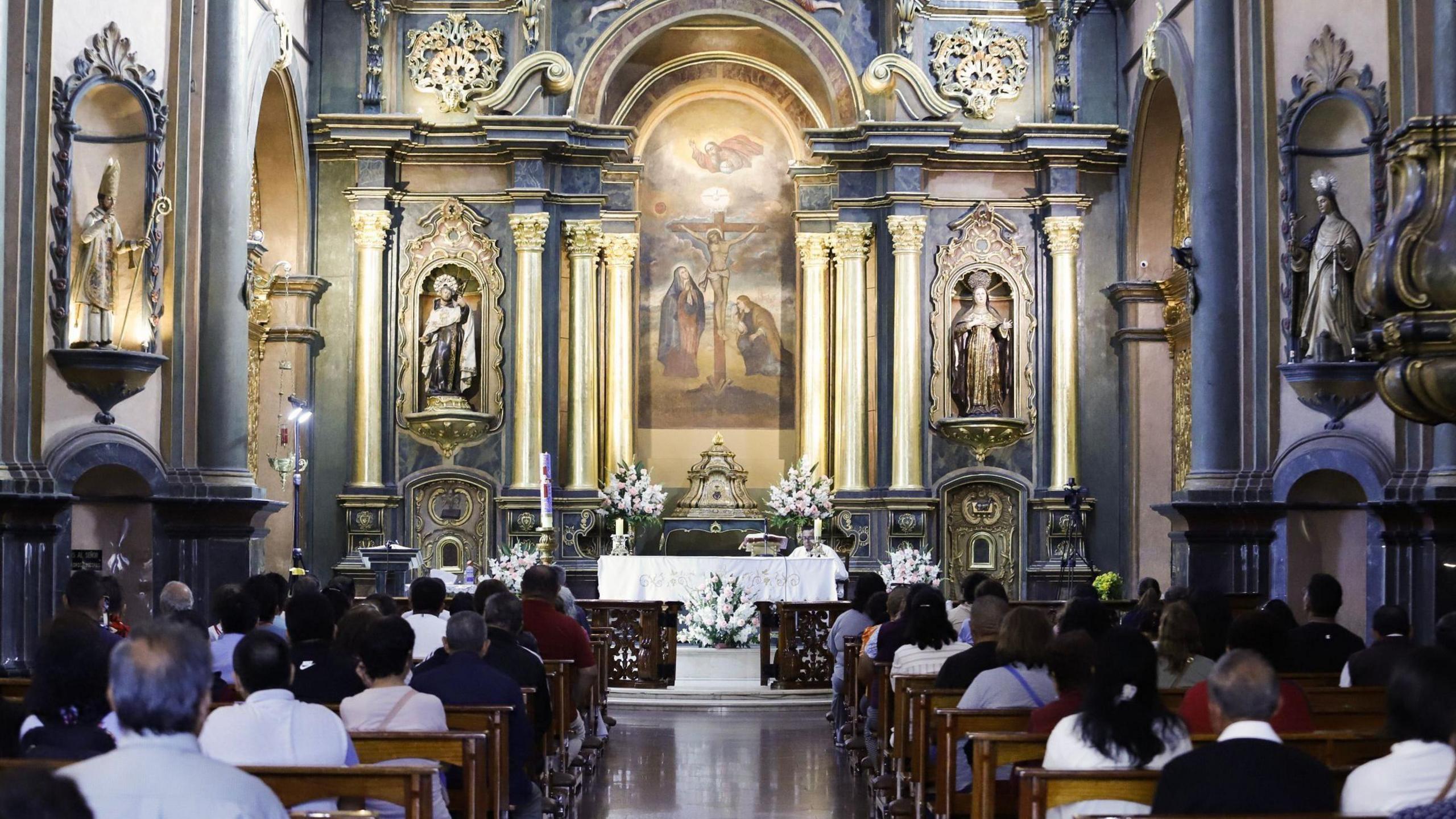 This picture is taken from the back of the church, and shows people sitting in wooden pews in mass, facing a painting of Jesus Christ on the cross.
