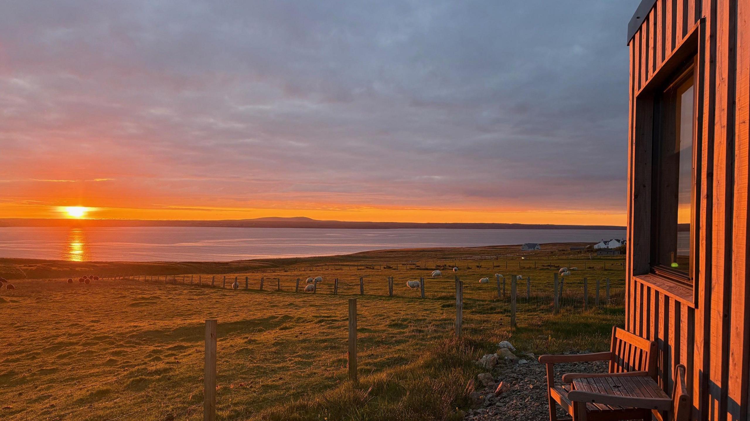 Sunset, with the sea in distance, and fields of sheep and the sun's glow against a wooden building.