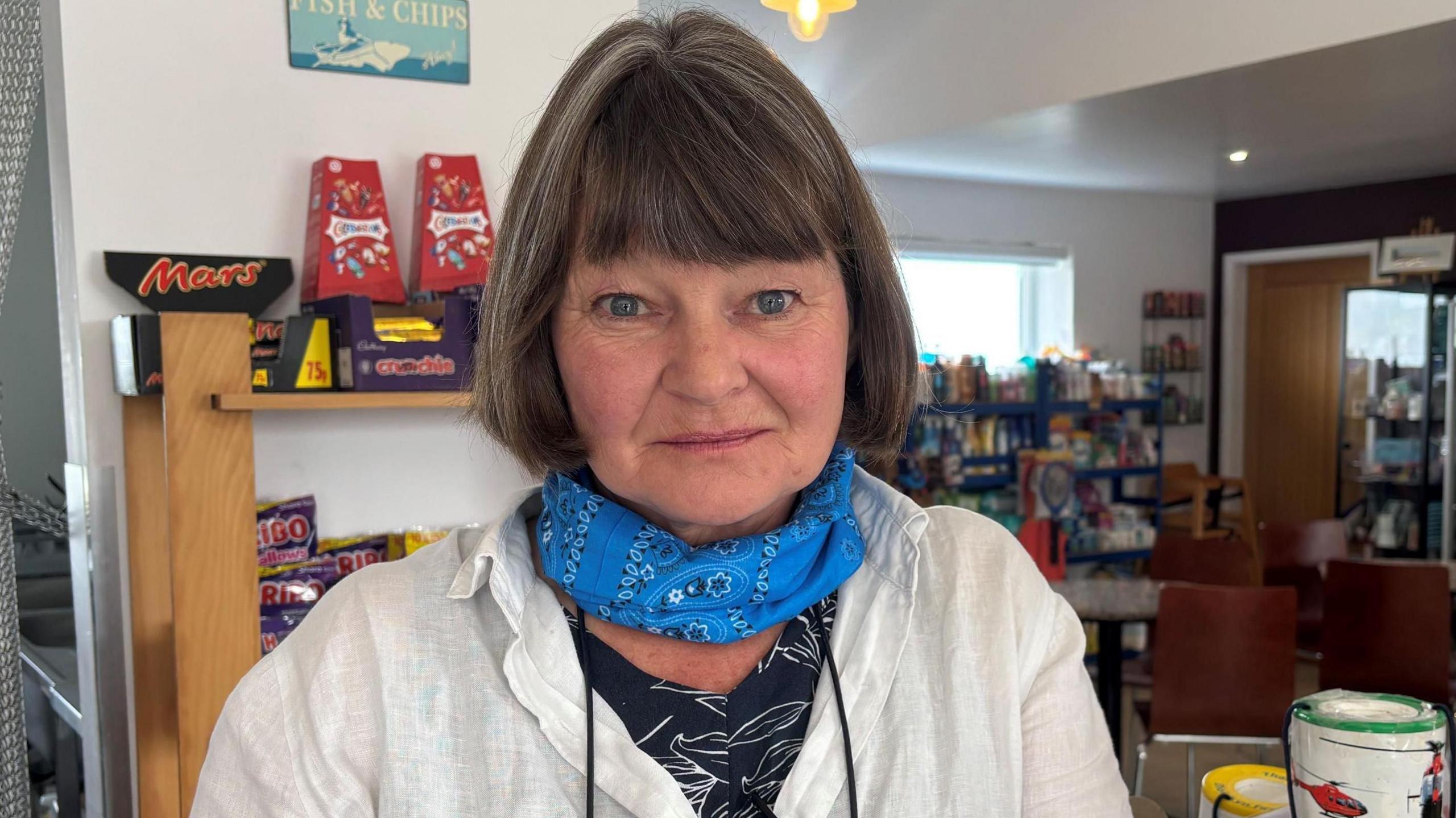 Close up image of Joanna smiling looking at the camera. She is stood in her campsite shop. She is wearing a cream linen shirt, a black floral top and a blue bandana around her neck. Joanna has short brown hair with a fringe. It is a head and shoulders shot. Behind her you can see shelves with sweets on.