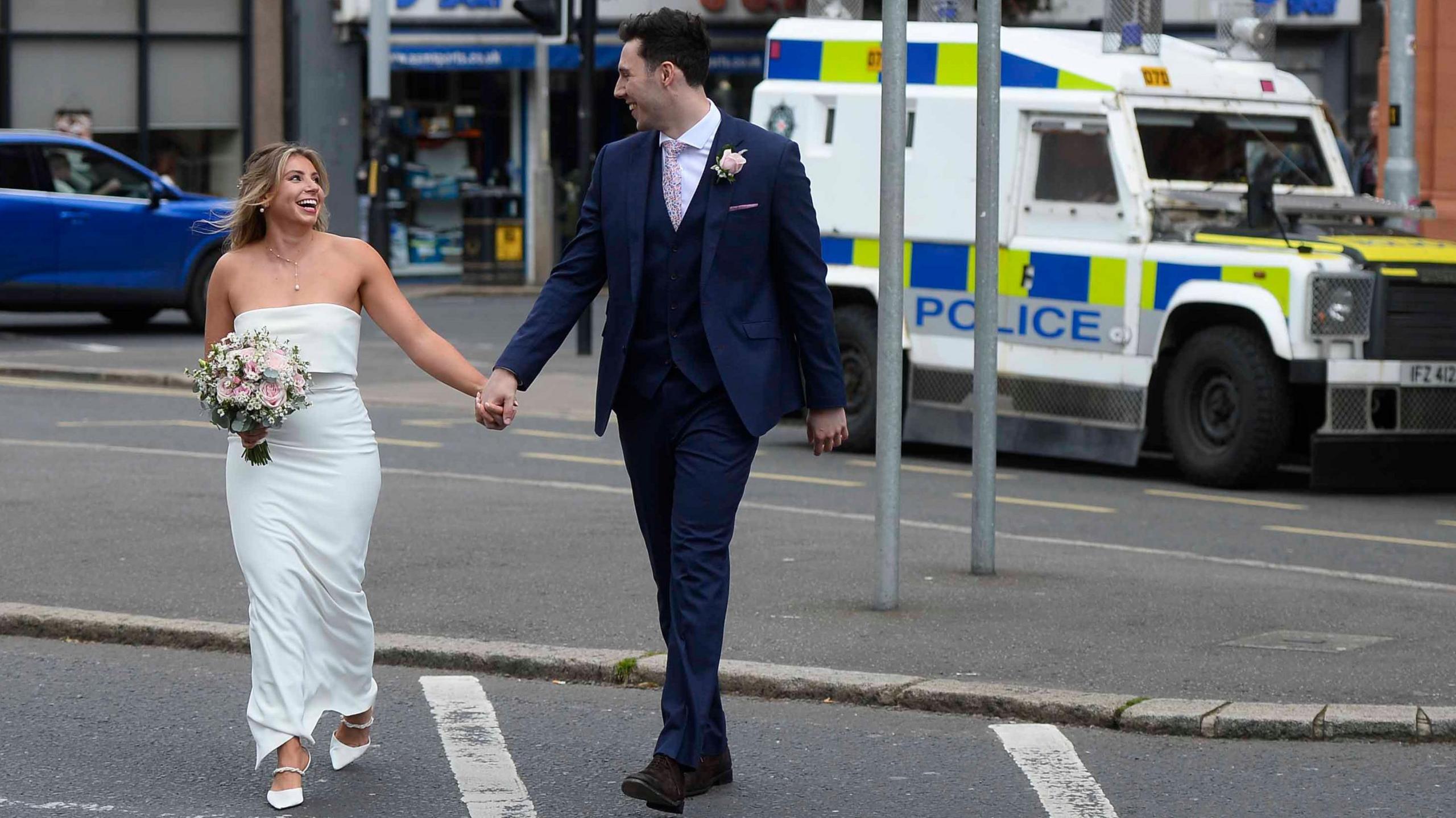 A bridge and groom hold hands with a police car in the background