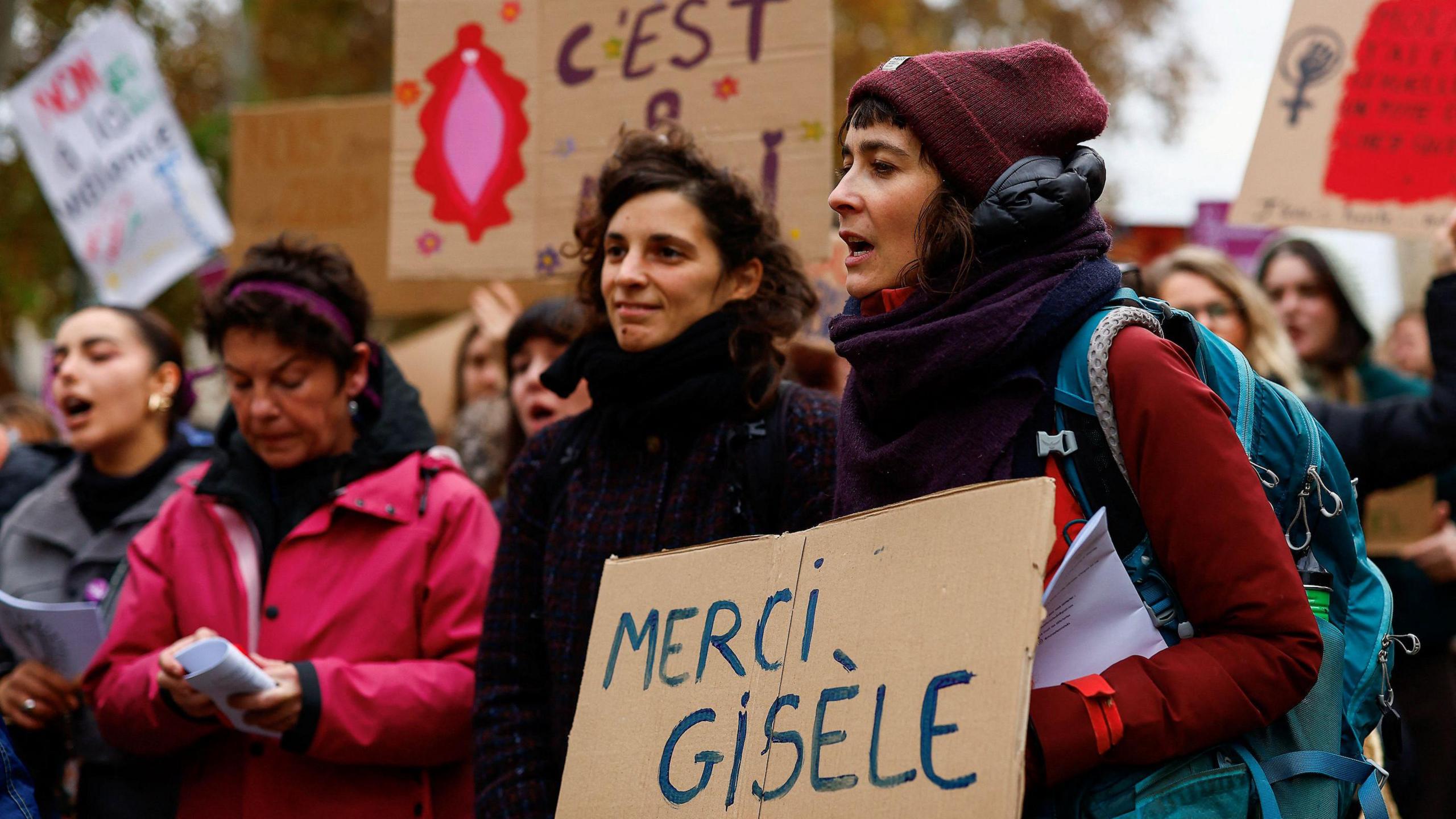 Women demonstrate outside the courthouse in Avignon, holding placards in support of Gisèle Pelicot. One at the front of the image holds a cardboard handwritten sign which says in French: "Thank you, Gisèle"