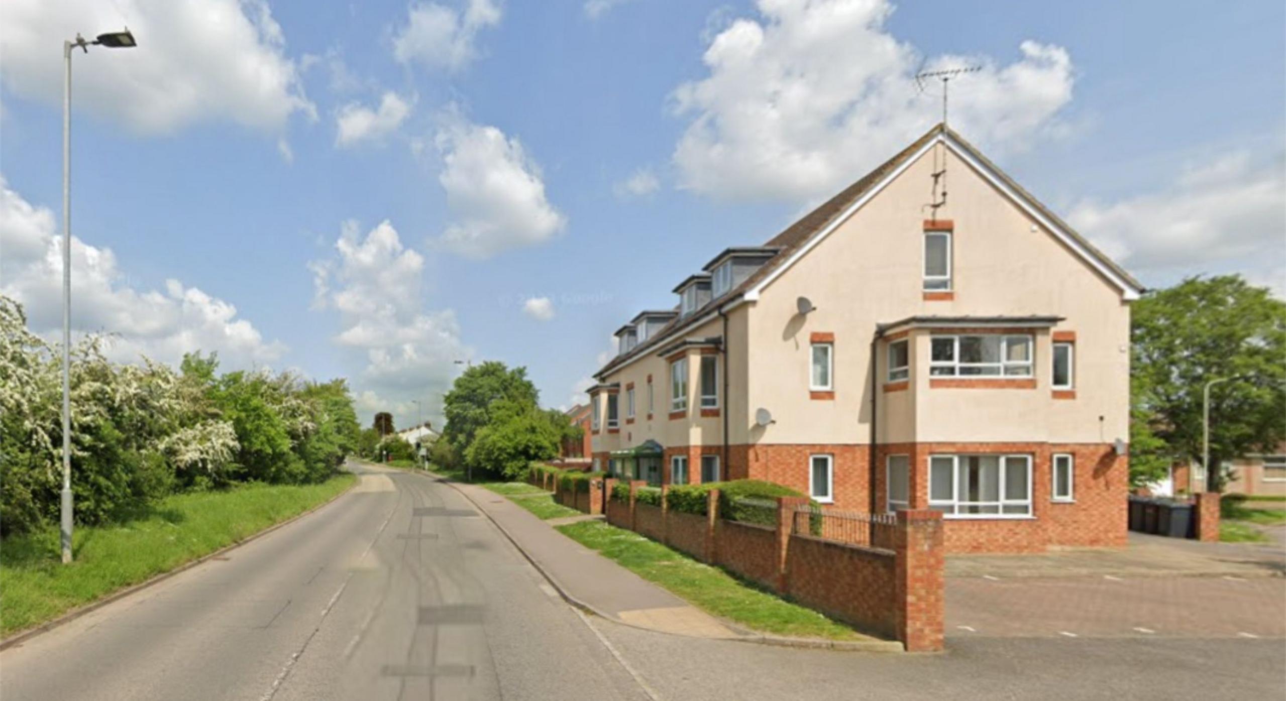 Village street with trees on one side and a two-storey modern residential building on the other. The top half is in light-coloured brick and the bottom half in red brick. There is a drive to the side of the property and a low brick wall separating it from the pavement. There is a grass verge between the wall and the pavement.
