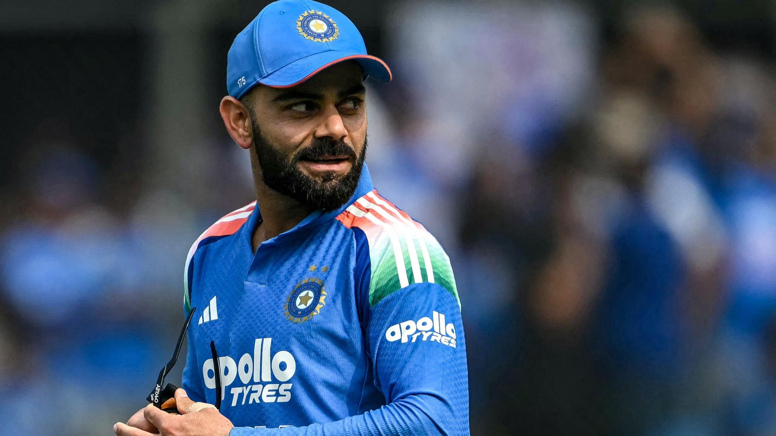 Virat Kohli looks behind, while polishing his sun glasses. He is wearing an India shirt and cap