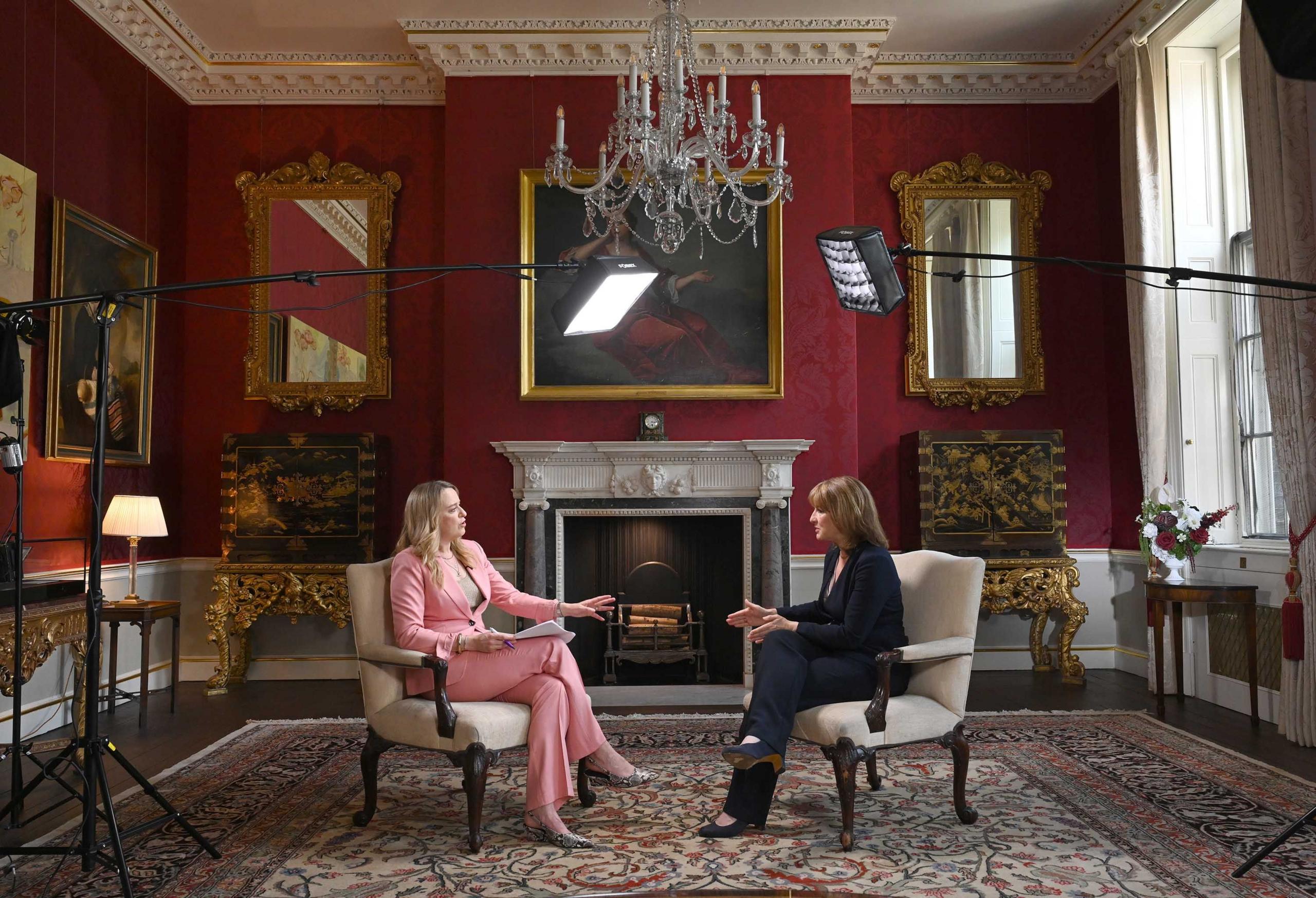Rachel Reeves on the right, in a blue suit, being interviewed by Laura Kuenssberg on the left, in a pink suit, in a grand room underneath a large chandelier
