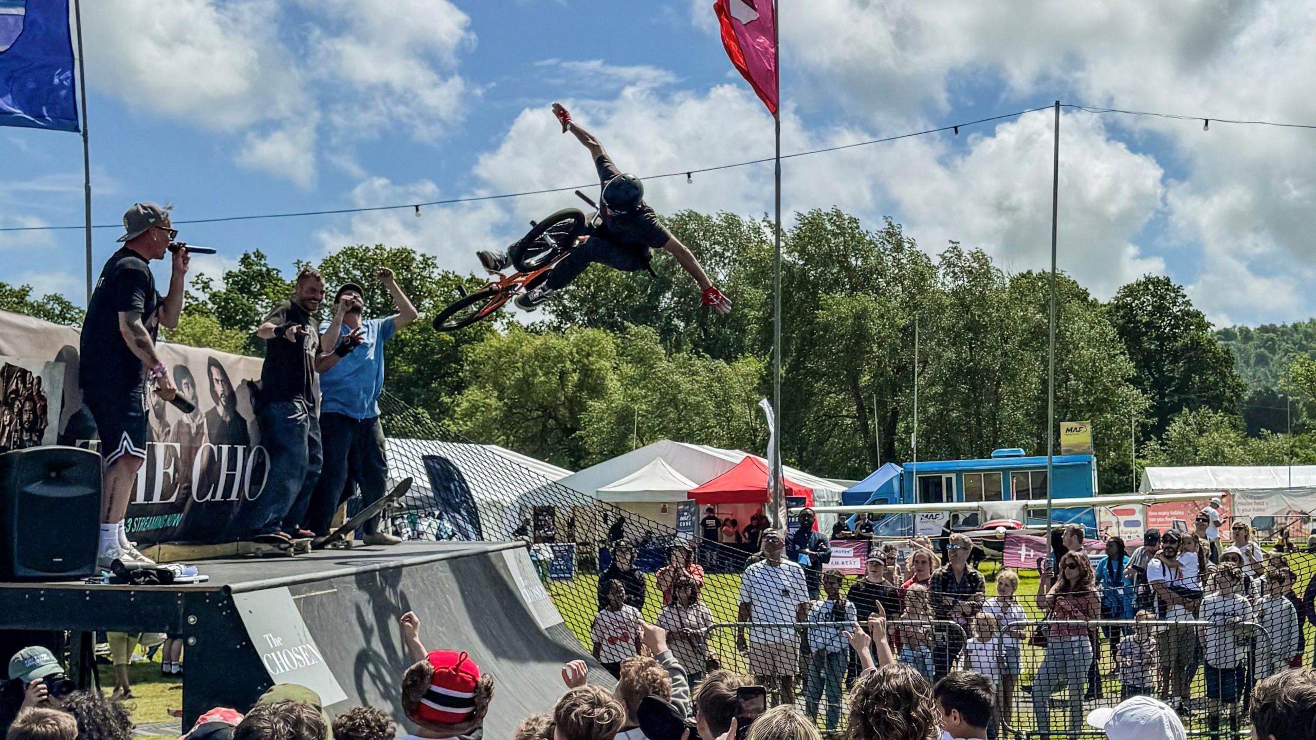 A BMXer performing a no hander in the air off a quarter pipe at an event. A crowd of people watch while announcers with microphones stand on top of the quarter pipe.