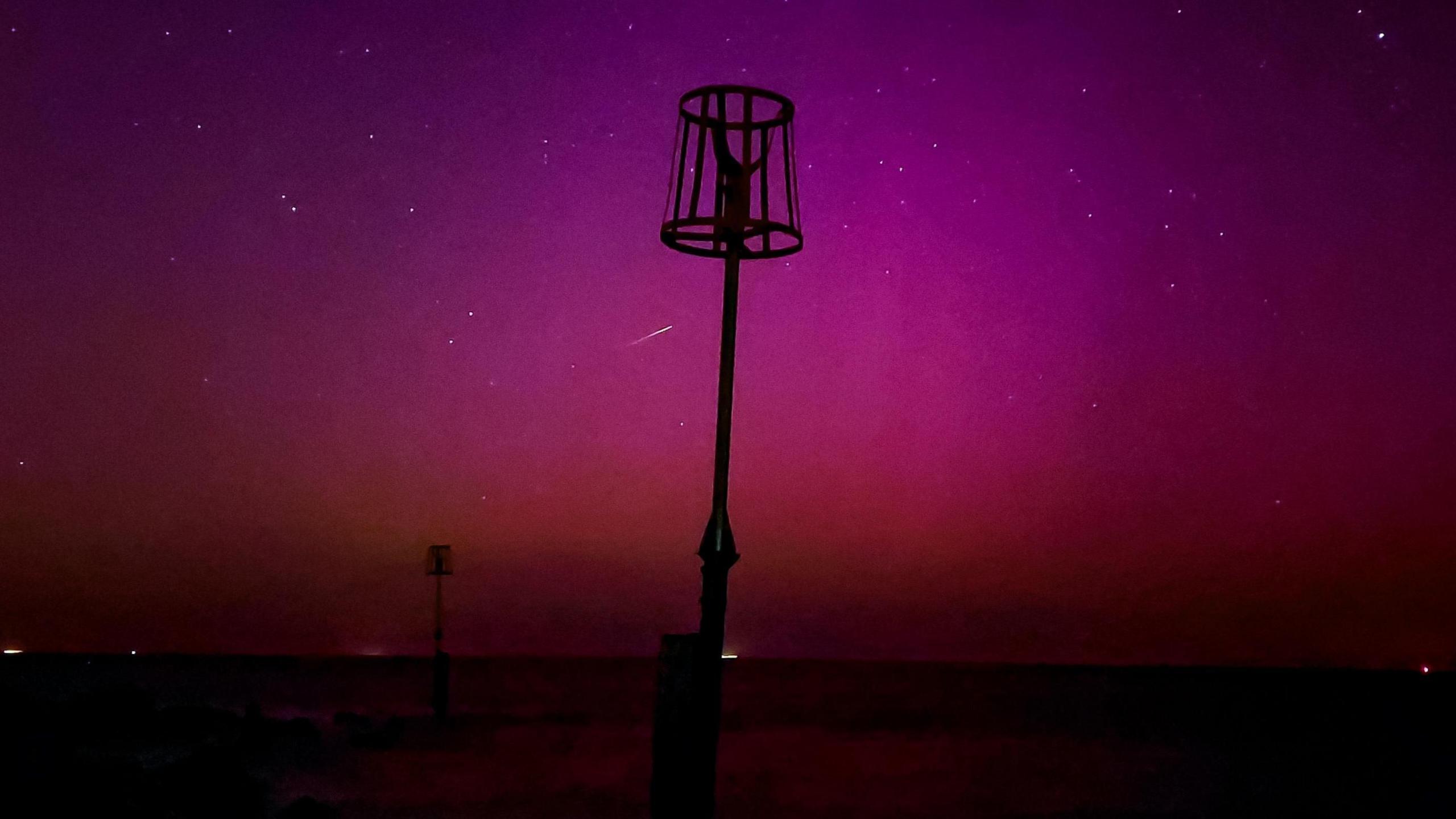 silhouette of the coastline and dark purples and reds indicating the aurora.  Few white dots of stars and a shooting star shown as a streak of white light