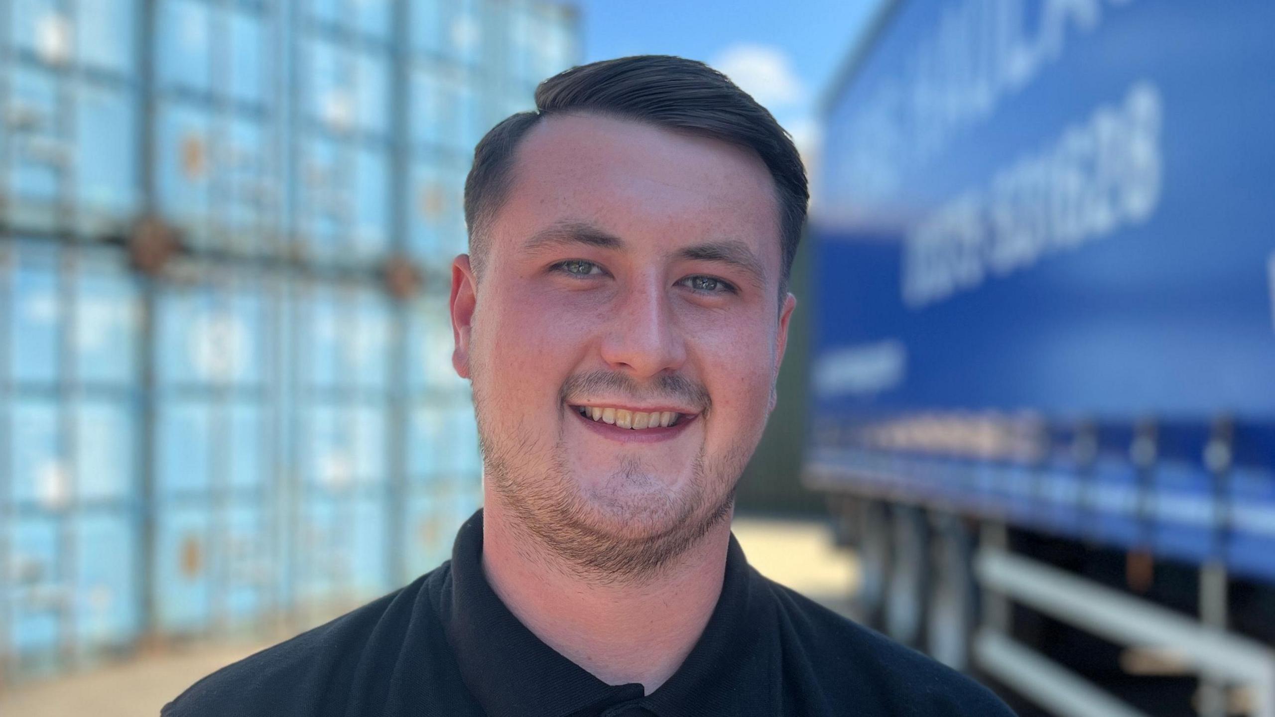 Frankie Stonebrook has short dark hair and is smiling while wearing a black polo shirt. He is standing in front of large blue containers and a blue lorry.
