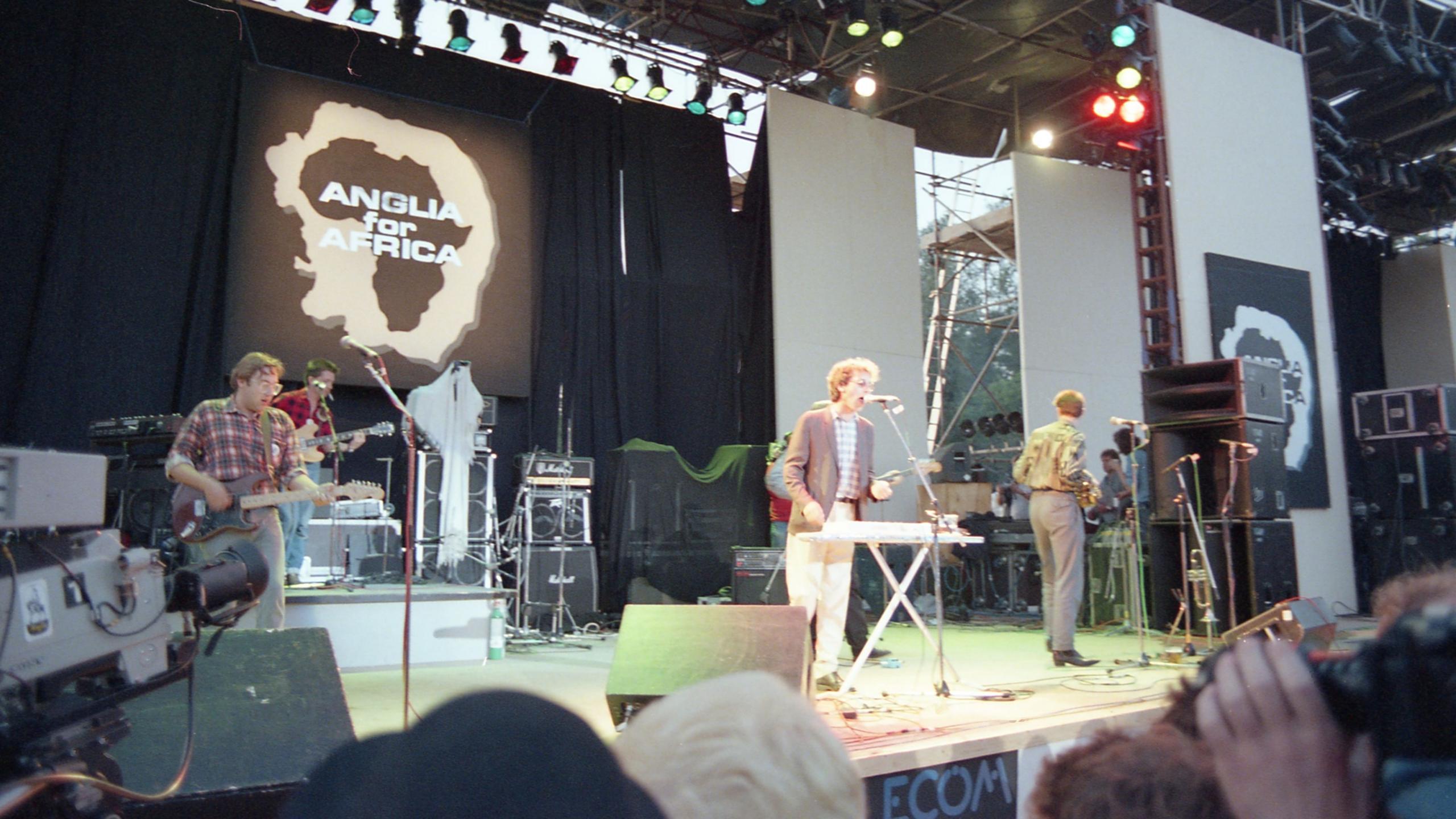 An outdoor stage with a large logo at the back of the stage reading 'Anglia for Africa', over a shape of Africa inside an outline shape of Norfolk and Suffolk. Performing on the stage is a pop group comprised of four young men, two playing guitars, one standing at a microphone while also playing a keyboard, and another man standing turned away from the camera. A few members of the audience are partly visible at the bottom right and bottom centre of the image, and a television camera pointing at the stage is visible bottom left.
