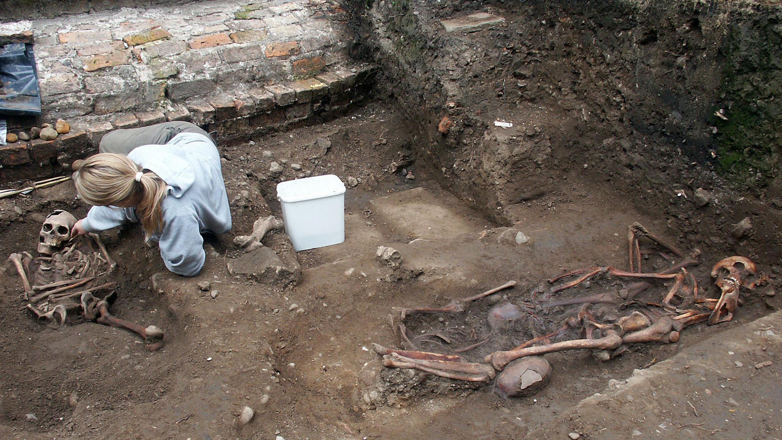 Skeletal remains lying in the soil bordered by a brick wall. A woman is on the ground next to one of the skeletons and there is a white bucket at her side. The woman has long blonde hair tied in a ponytail and is wearing a blue jumper and grey trousers.