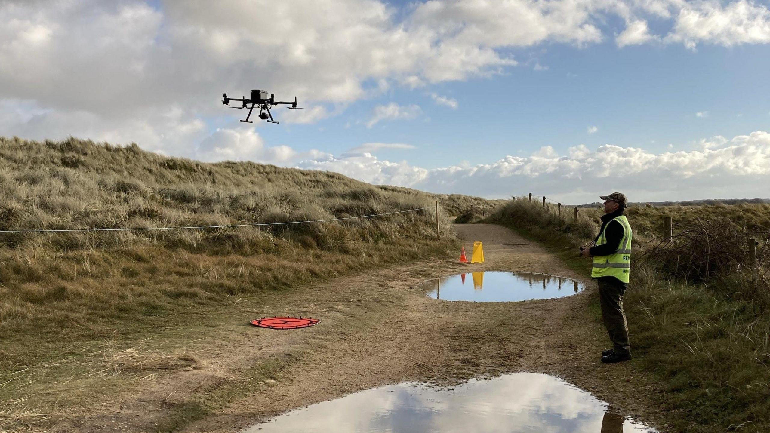 A drone flies in the air at a low level over a stretch of fenced off dune, with a male operator - dressed in a high-vis vest and baseball hat - standing nearby on a path covered in puddles and with the drone control in his hands.