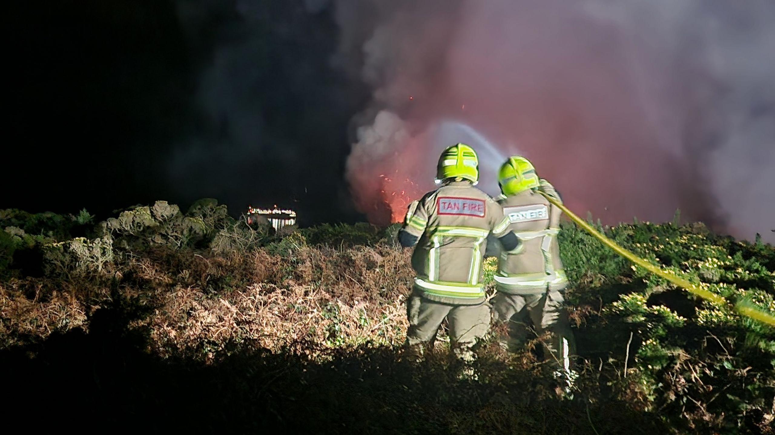 Two firefighters wearing helmets are seen dousing a fire on a hillside, with large pink and red flames ahead of them