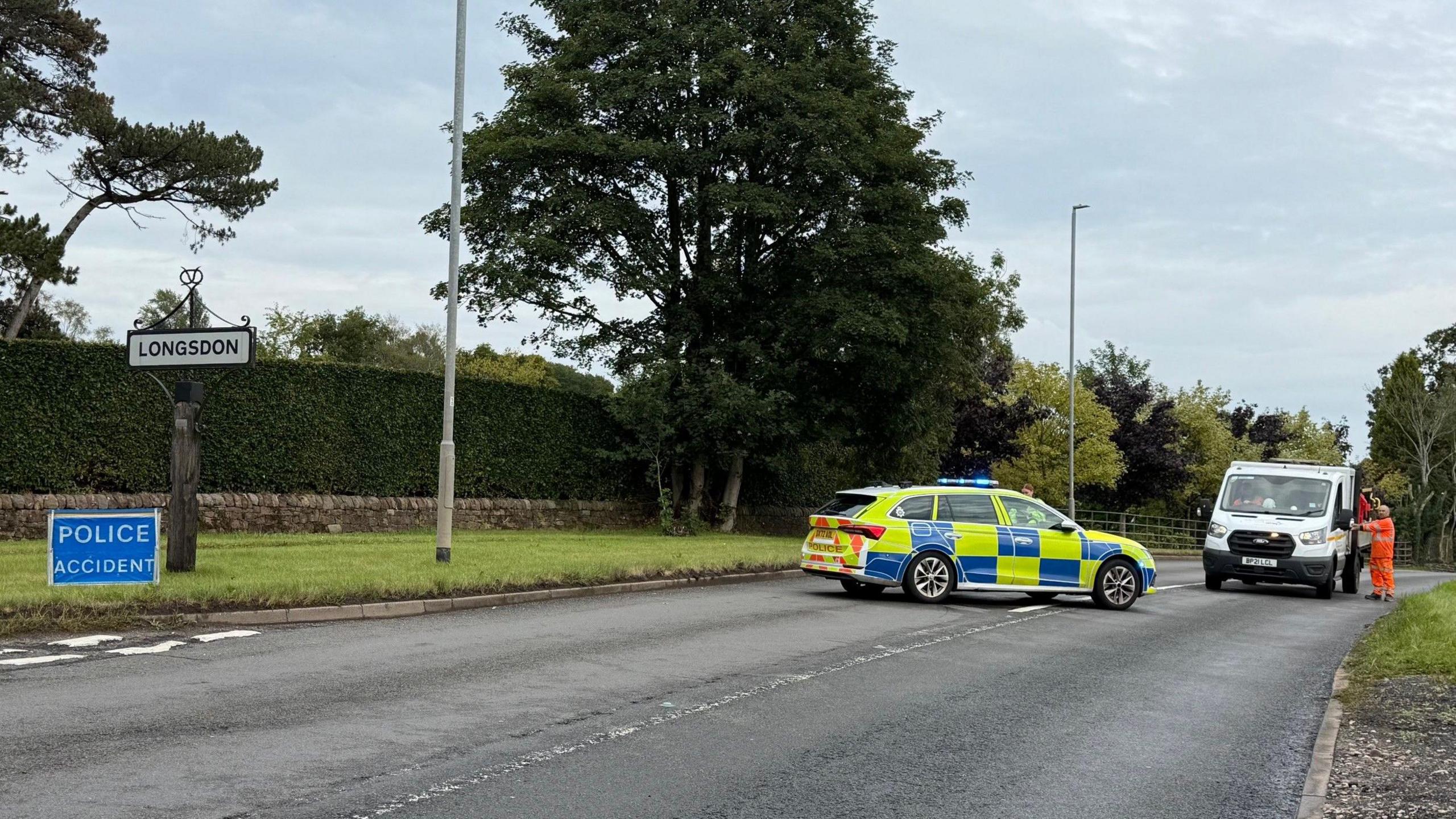 A police car is parked across a road, with a highways van parked alongside it. There is a blue sign on the grass verge indicating a road closure as a result of an incident.