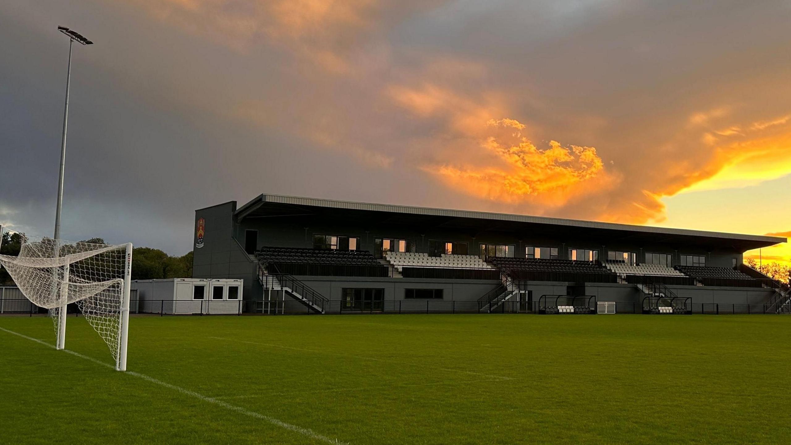 Cambridge City FC ground at Sawston. A grey football stadium stand and the above is a yellow/orange sunset. There is also a goal with the net folded up at the end of the pitch which runs in front of the stand