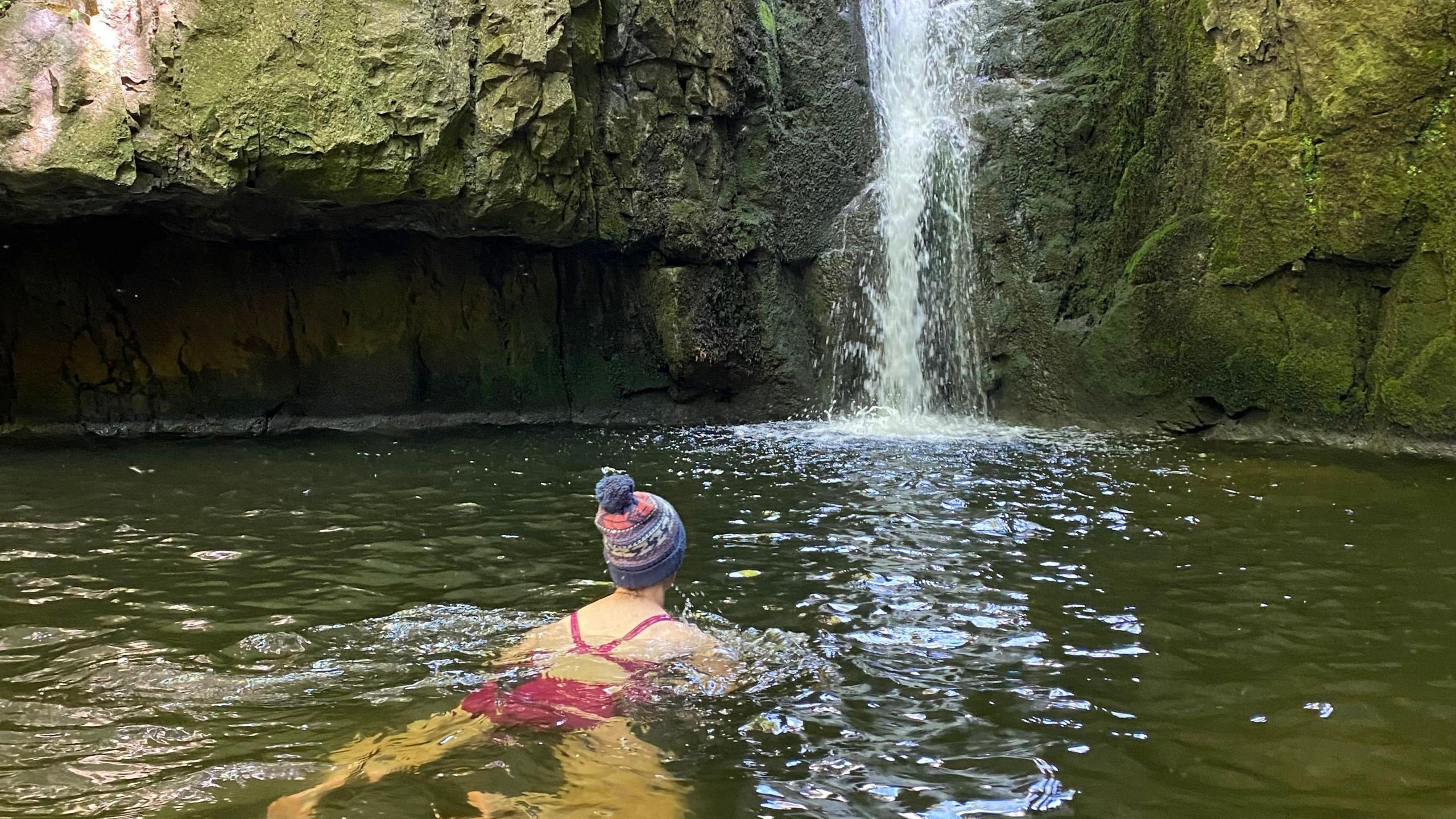 Bex Hind swims in a pool of water in a beauty spot with a waterfall. She is wearing a pink swimsuit and has a bobble hat.