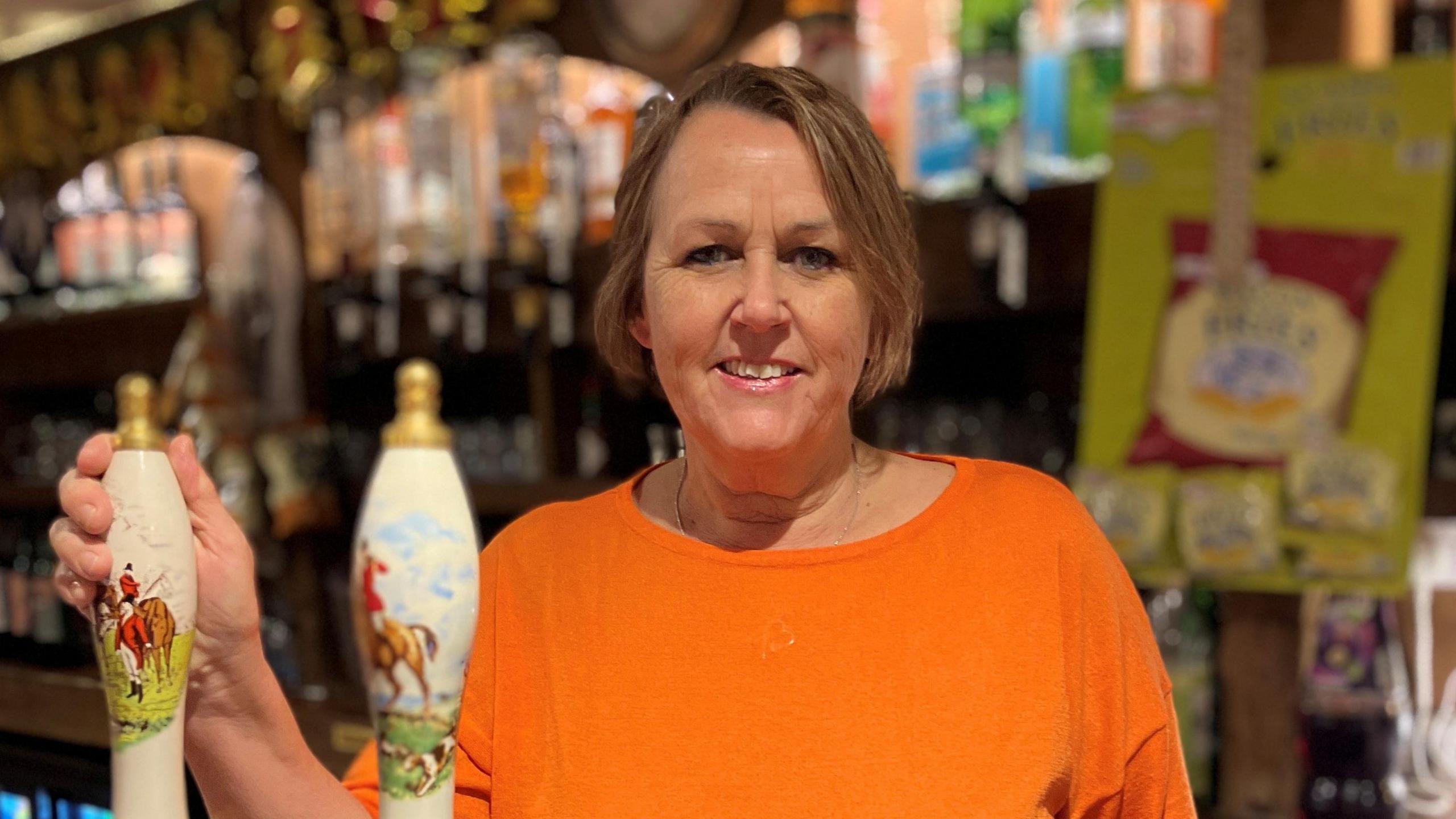 The manager of the pub, Maria Swanscott, standing at the bar of The Framsden Greyhound next to hand pumps. She is wearing an orange top and is smiling.