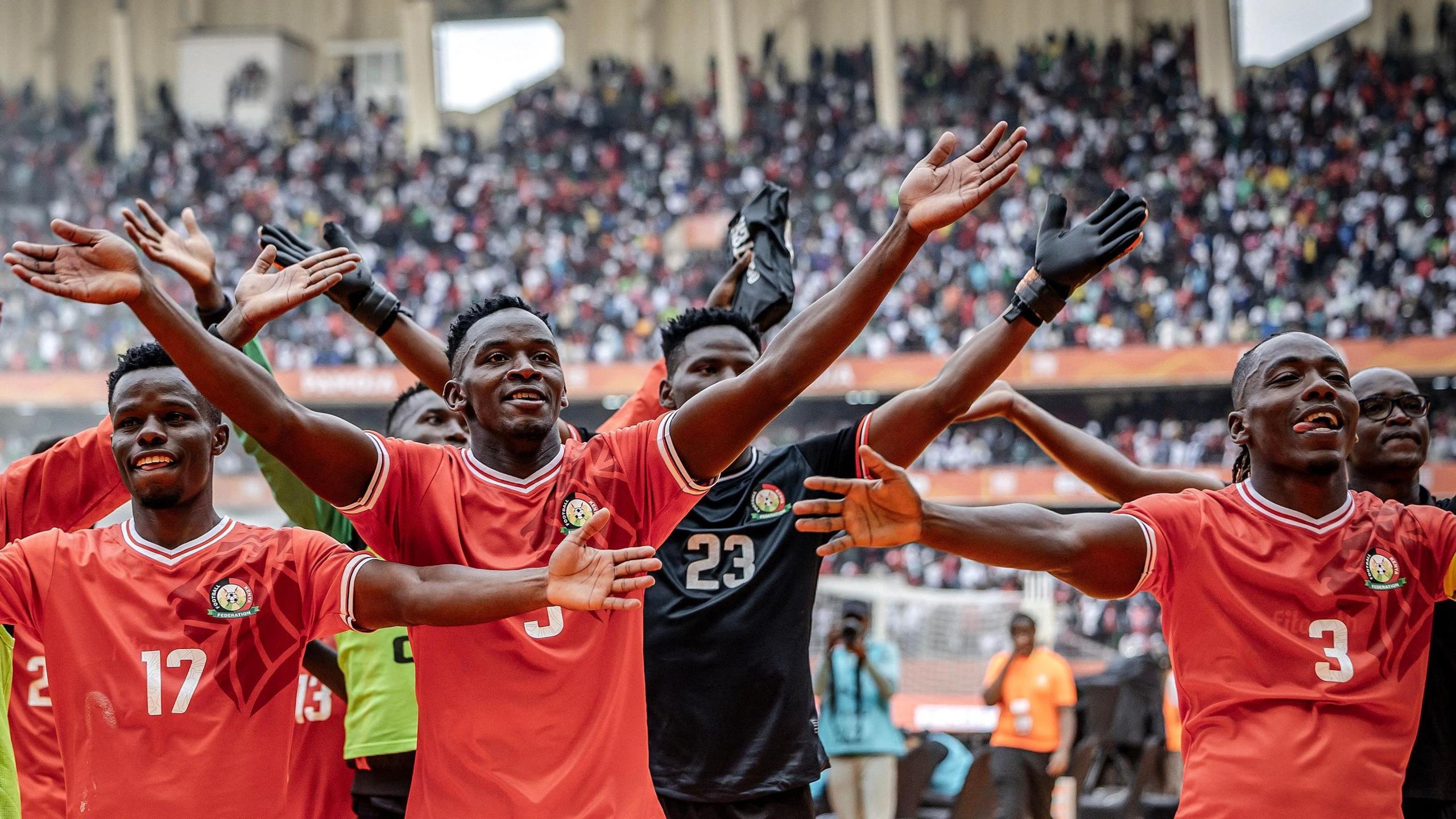 Kenya players in red shirts, as well as a goalkeeper in black, hold their arms up aloft in celebration after a football match while in the far distance fans are seen in packed stands