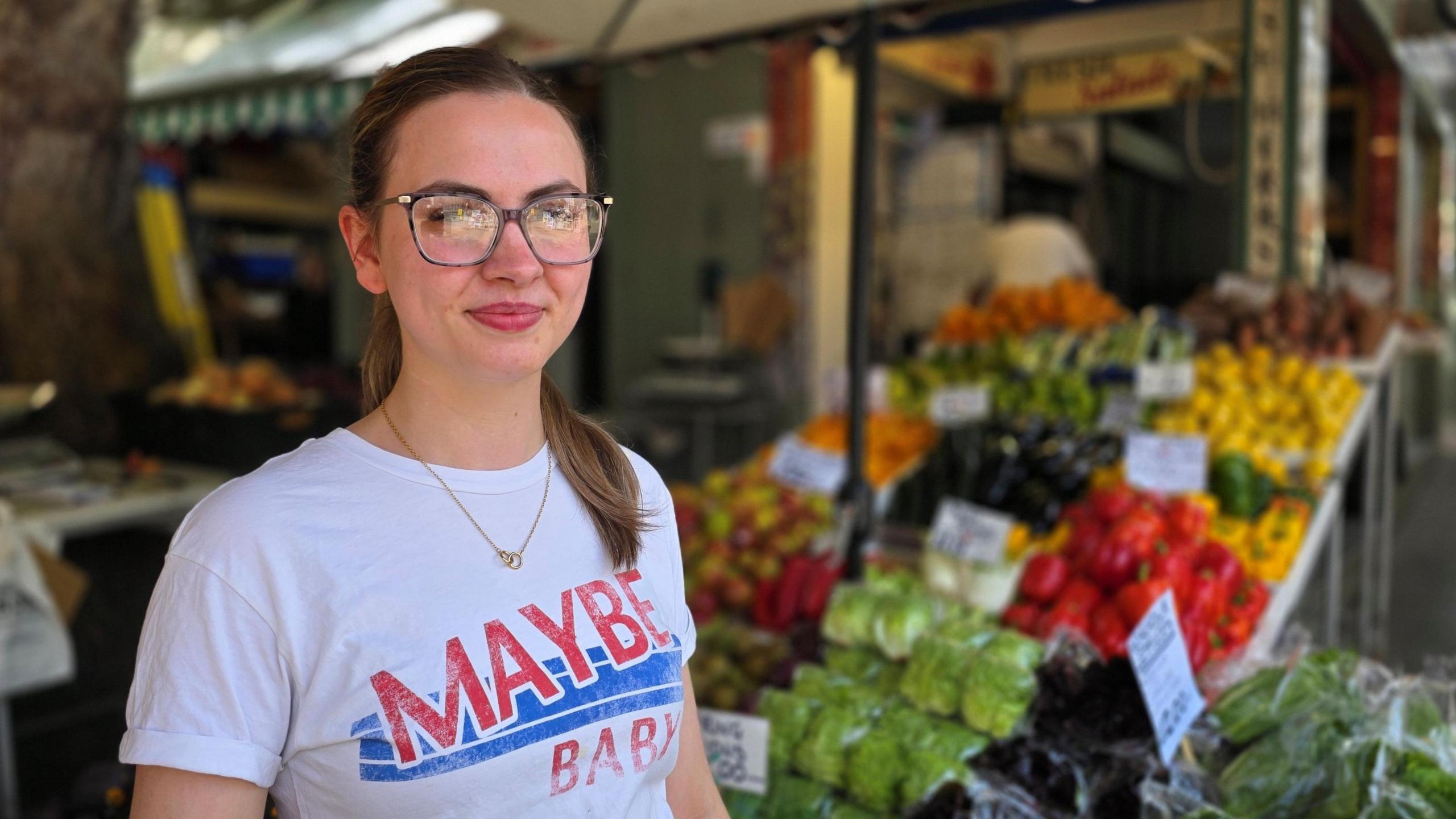 Billie Waring is wearing a white t-shirt with 'Maybe Baby' on the front. She has large framed glasses and we can see a selection of vegetables behind her.