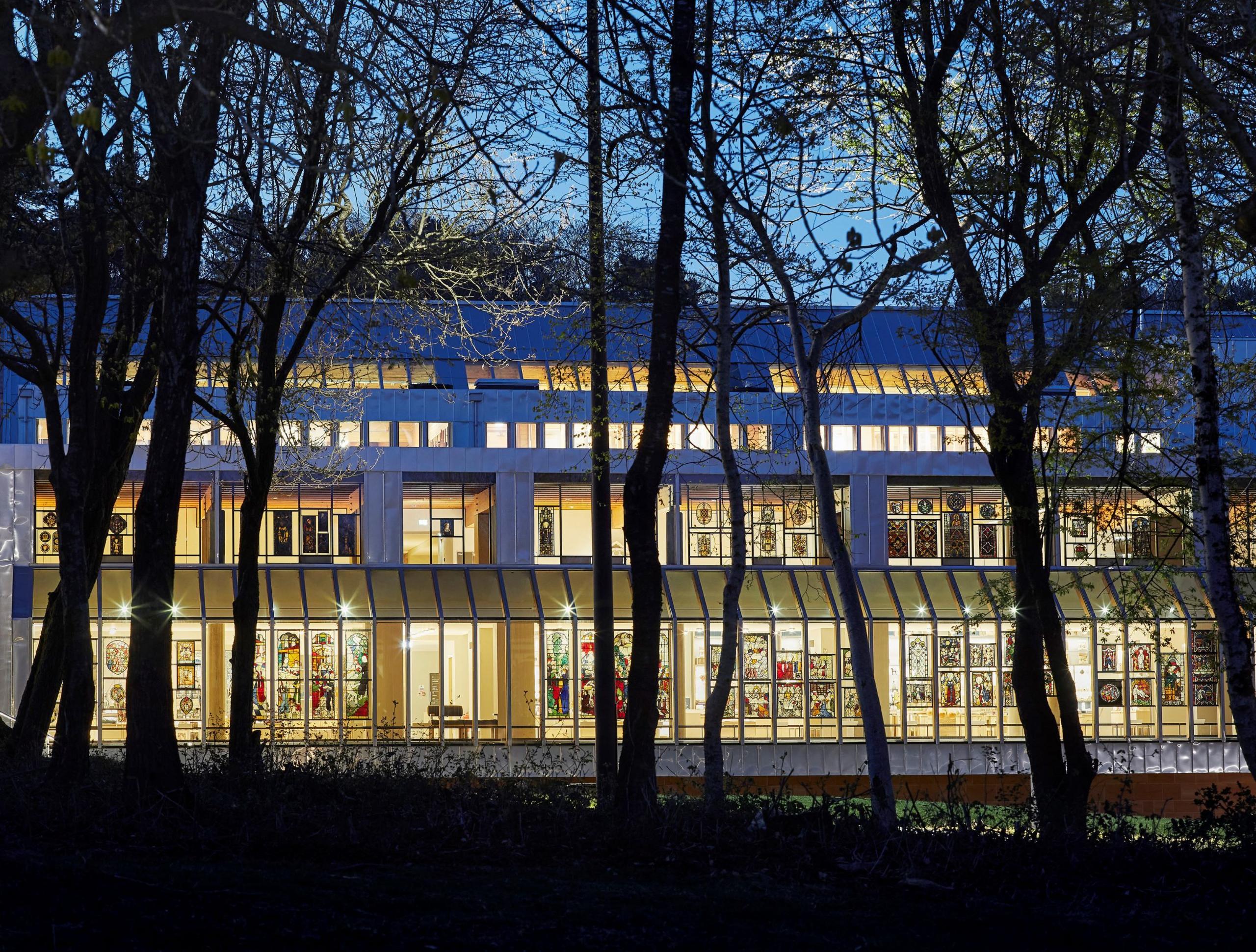 Another outside view, this time closer, looking into the lit-up gallery through a row of dark trees.