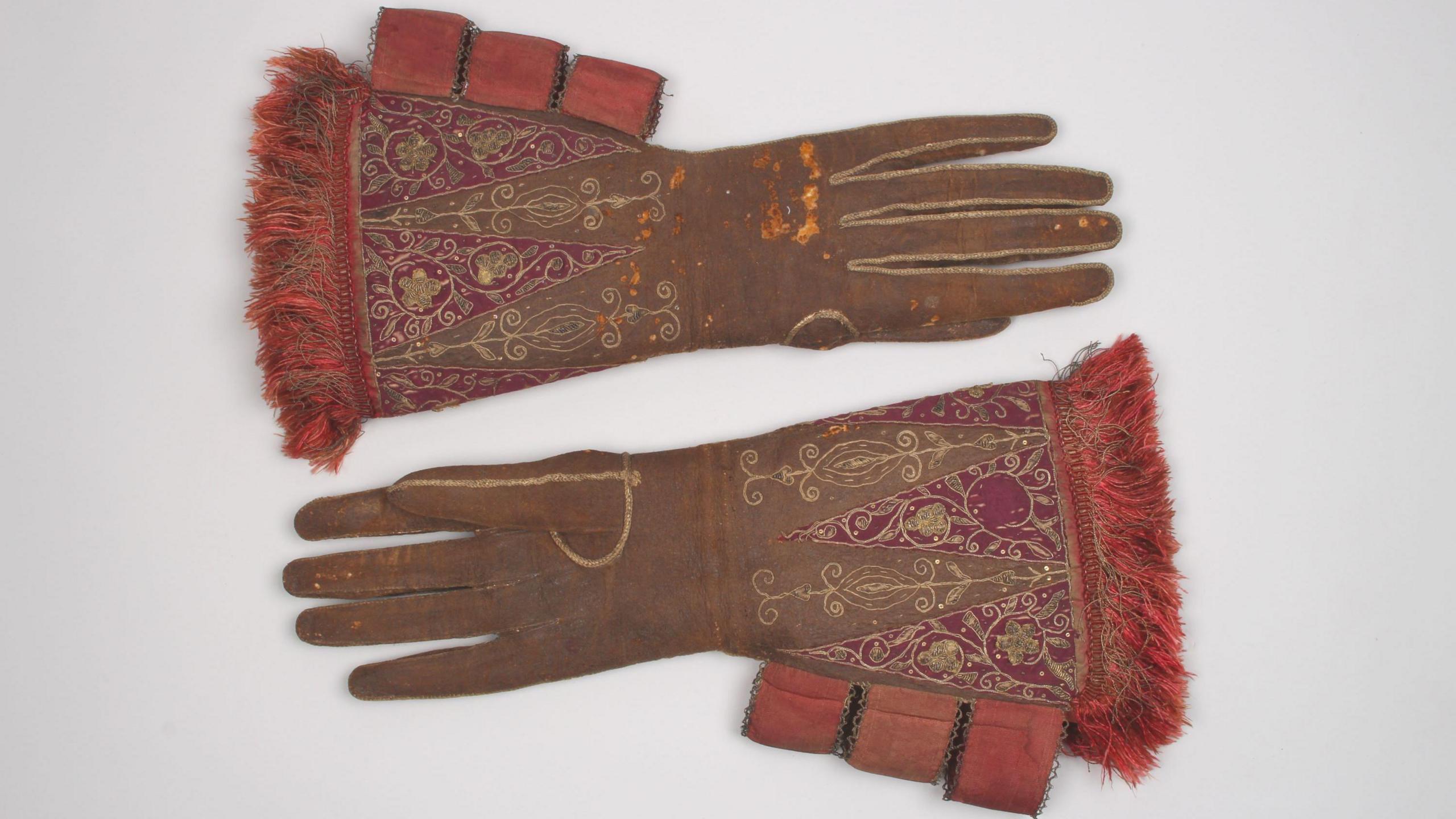 A pair of large brown leather gloves with red silk embroidery and gold embellishment, belonging to King James I, circa 1610s. They are on a grey background.