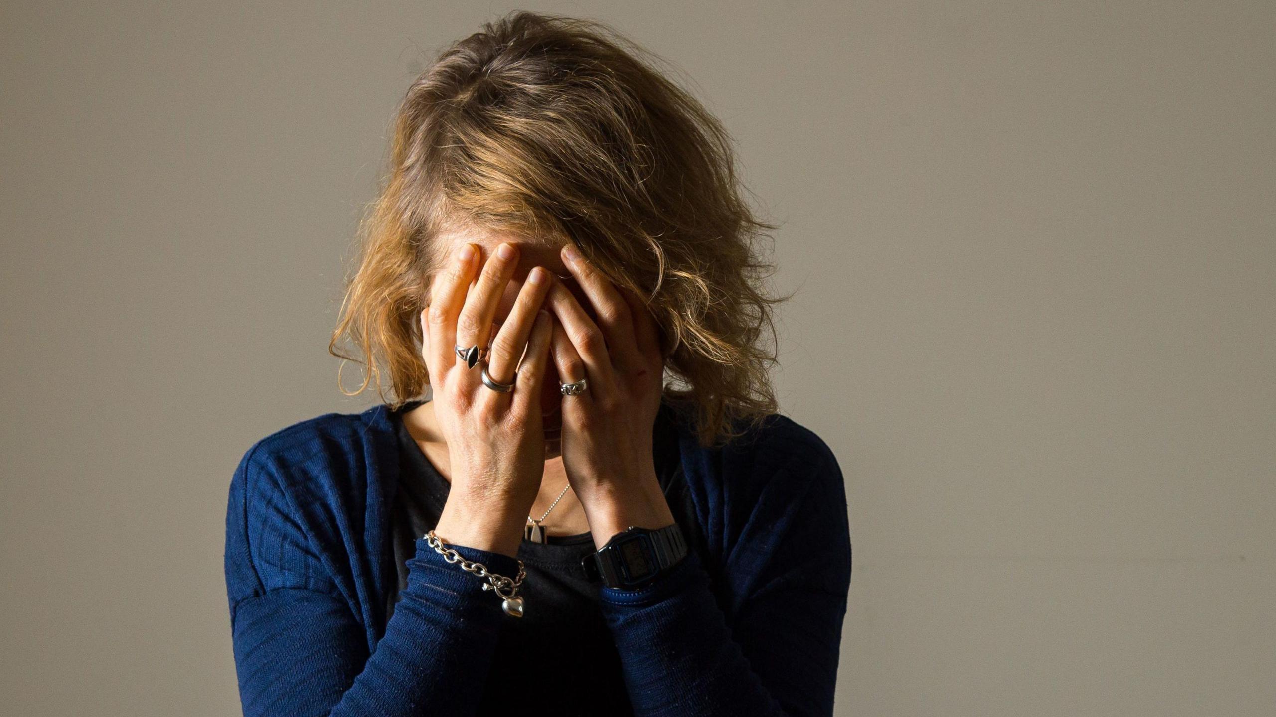 A woman in a blue top is stood facing the camera with her head in her hands. She has multiple rings on her fingers and a bracelet. 