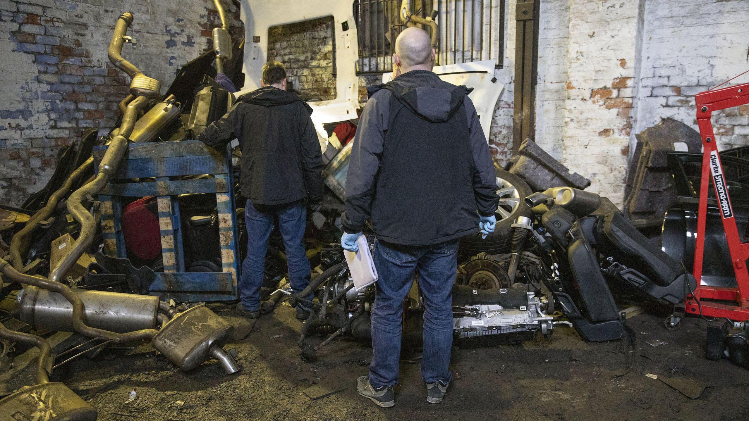 Two police detectives look on at a a pile of broken up car parts in a warehouse. 