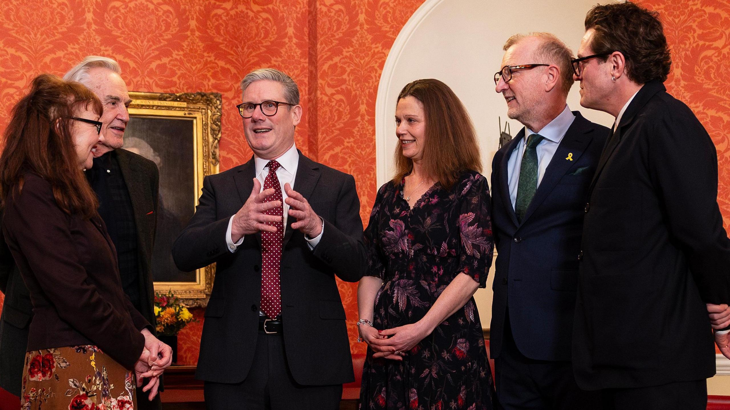 Keir Starmer stands surrounded by members of the Gavin & Stacey cast. Larry Lamb and Melanie Walters stand to his left hand side, while Mathew Horne and Steffan Rhodri stand to his right with a woman with dark hair wearing a floral dress. They are standing in a room with bright orange wallpaper and a portrait with a gilded frame. 