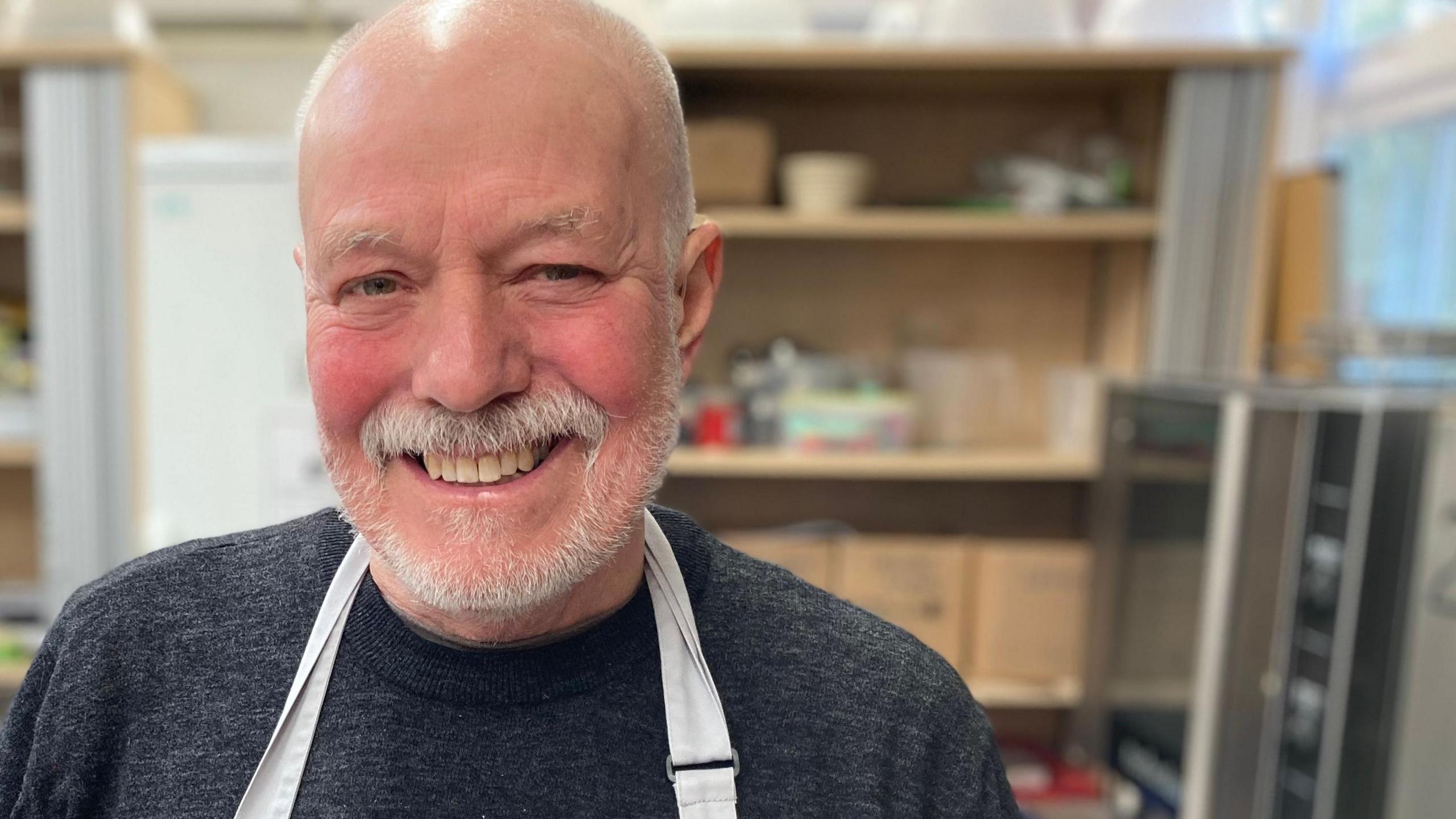 A man wearing an apron is pictured smiling in a kitchen. He had a white beard and is wearing a grey top.