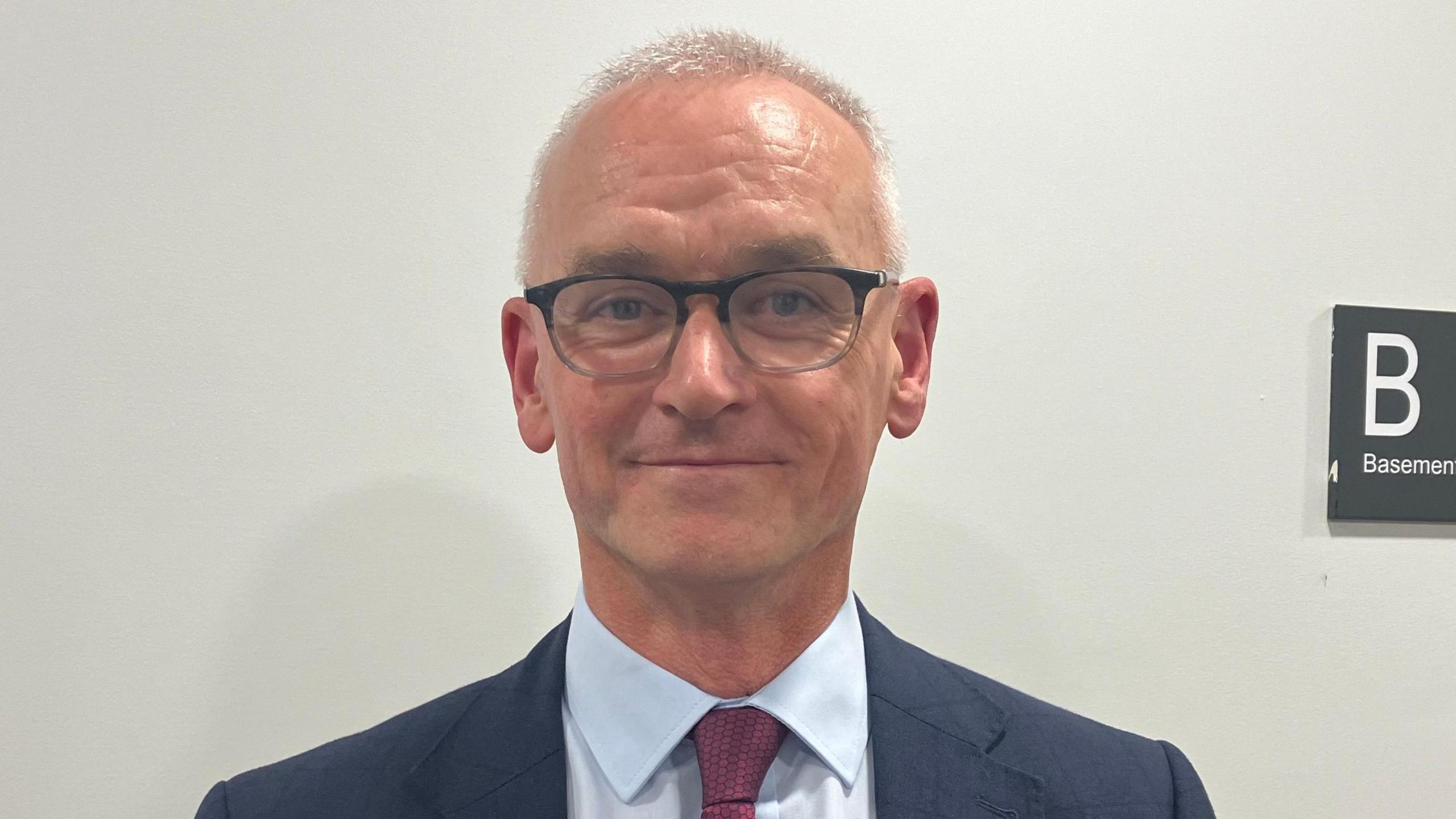 Alan Barrett is standing in front of a white wall. He wears glasses and is wearing a navy suit with a red tie and a light blue shirt. 