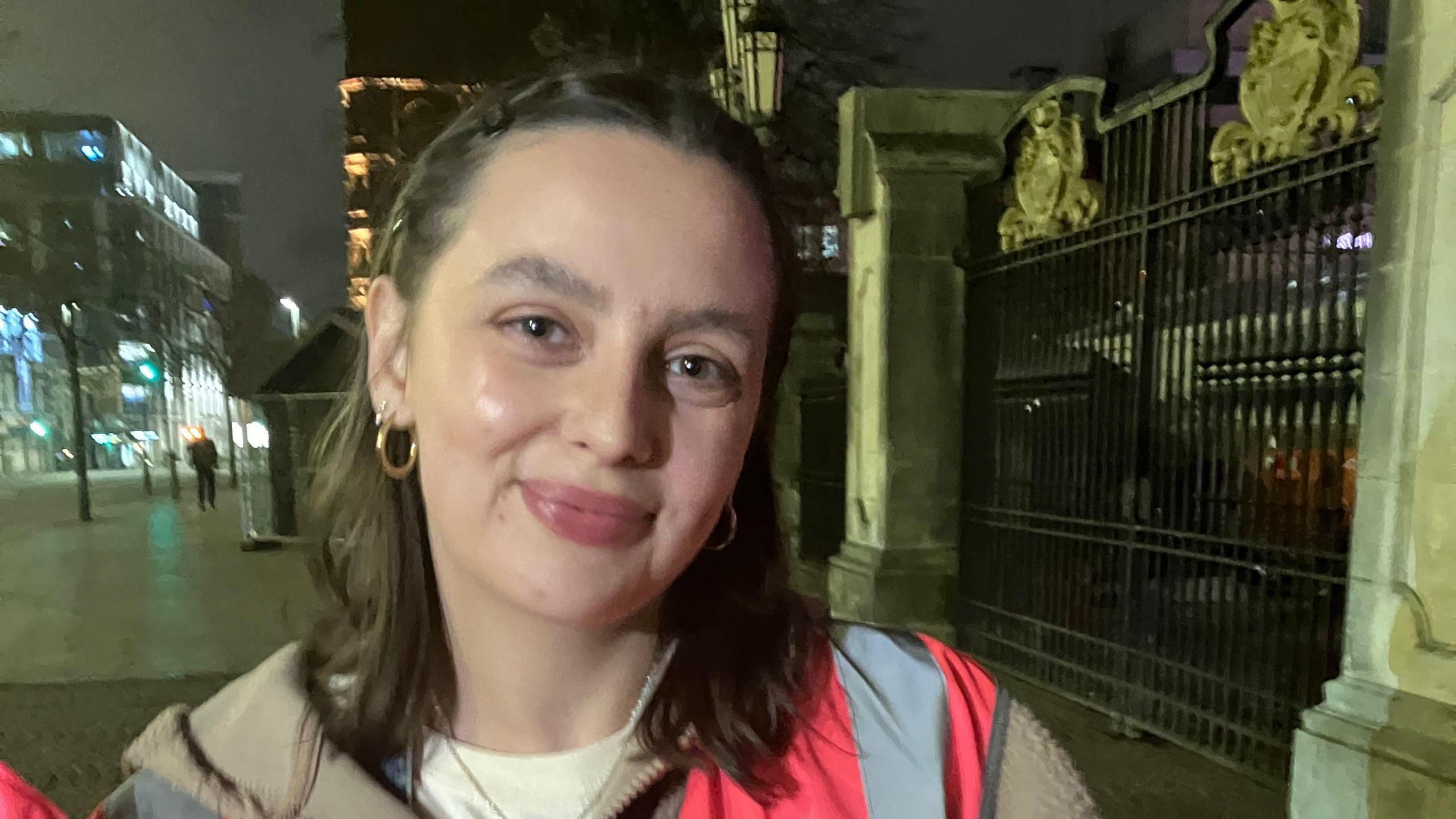 A girl with brown hair stands in front of city hall, she has on a brown jumper underneath a pink hi-viz. She has on golden earings