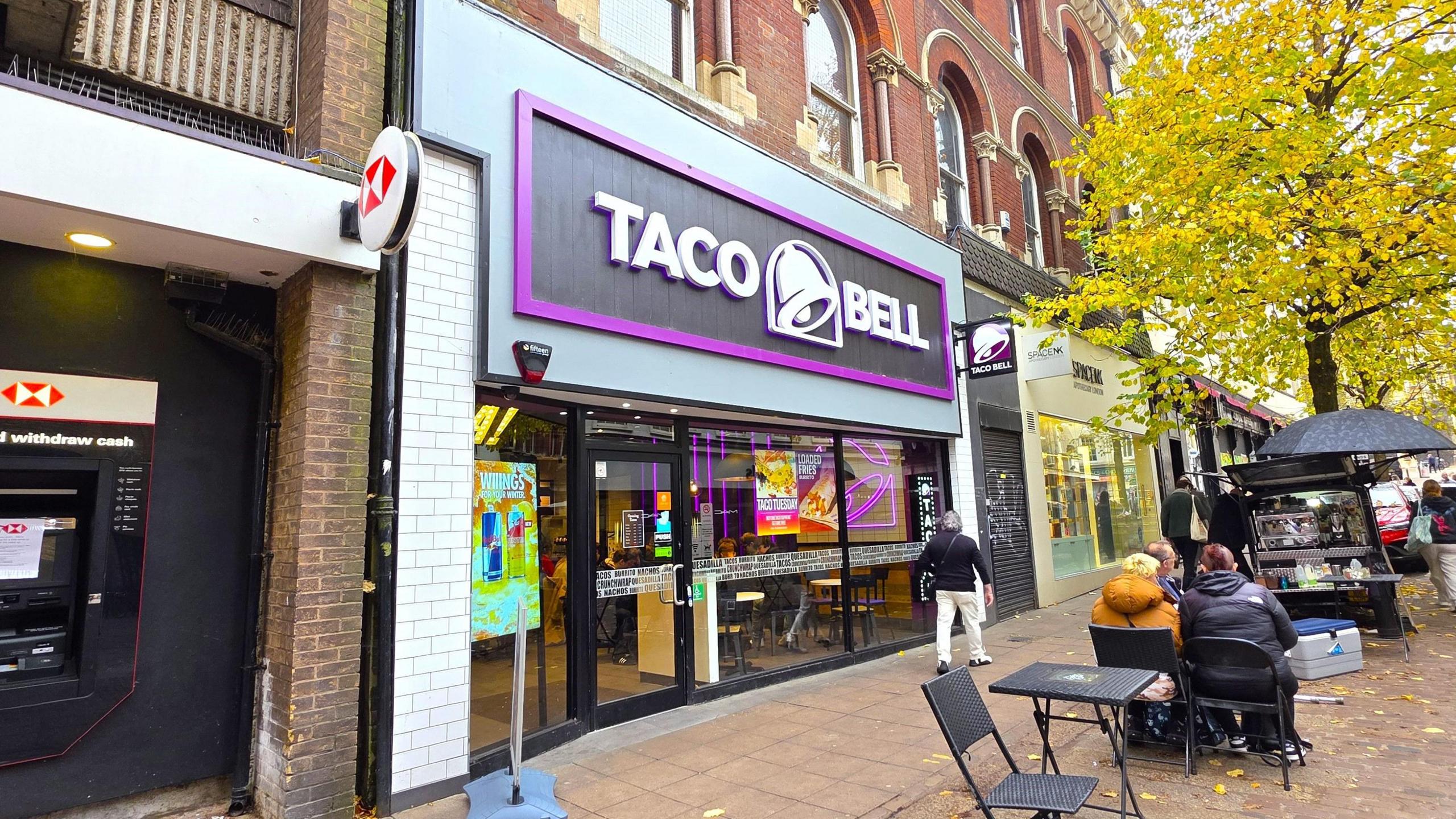 The exterior of Taco Bell. It is a small shopfront with a black and purple backing for the shop sign, which also features a bell. Outside we can see a couple of tables and chairs, and people sitting or walking nearby.