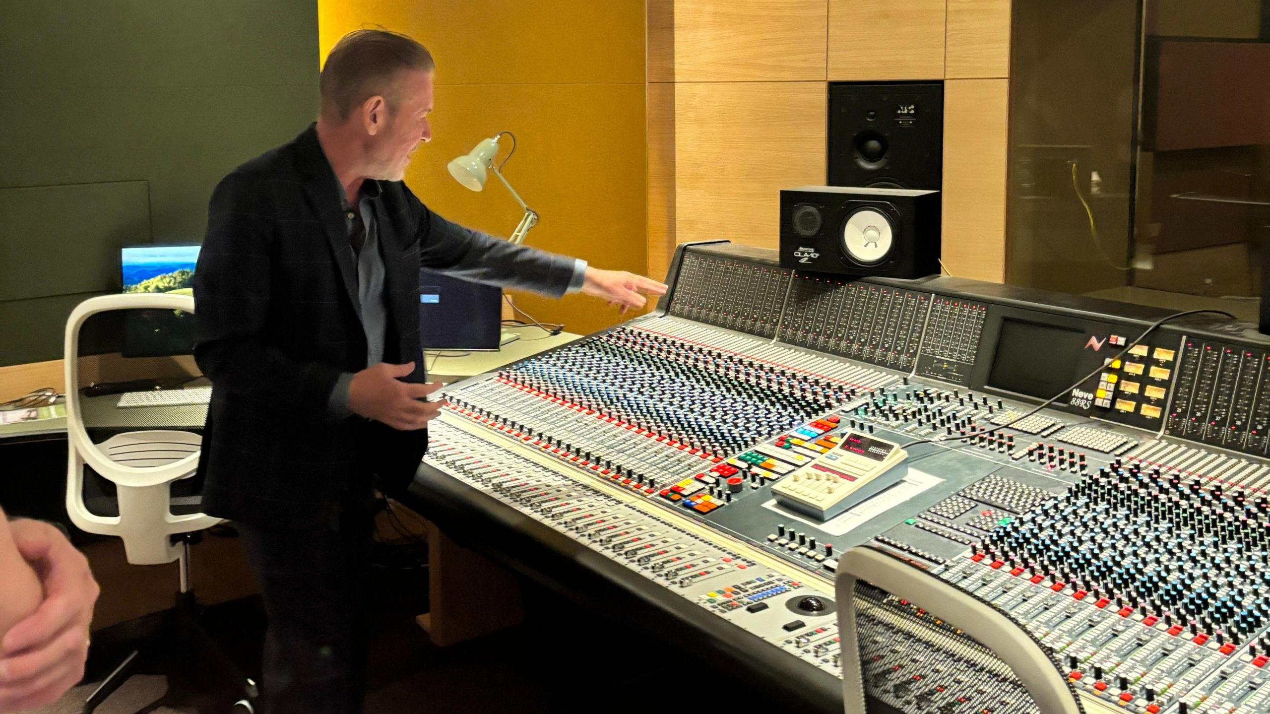 Steve Lillywhite, wearing a blue shirt and a dark suit, looks over a studio mixing desk with dozens of faders and channels.