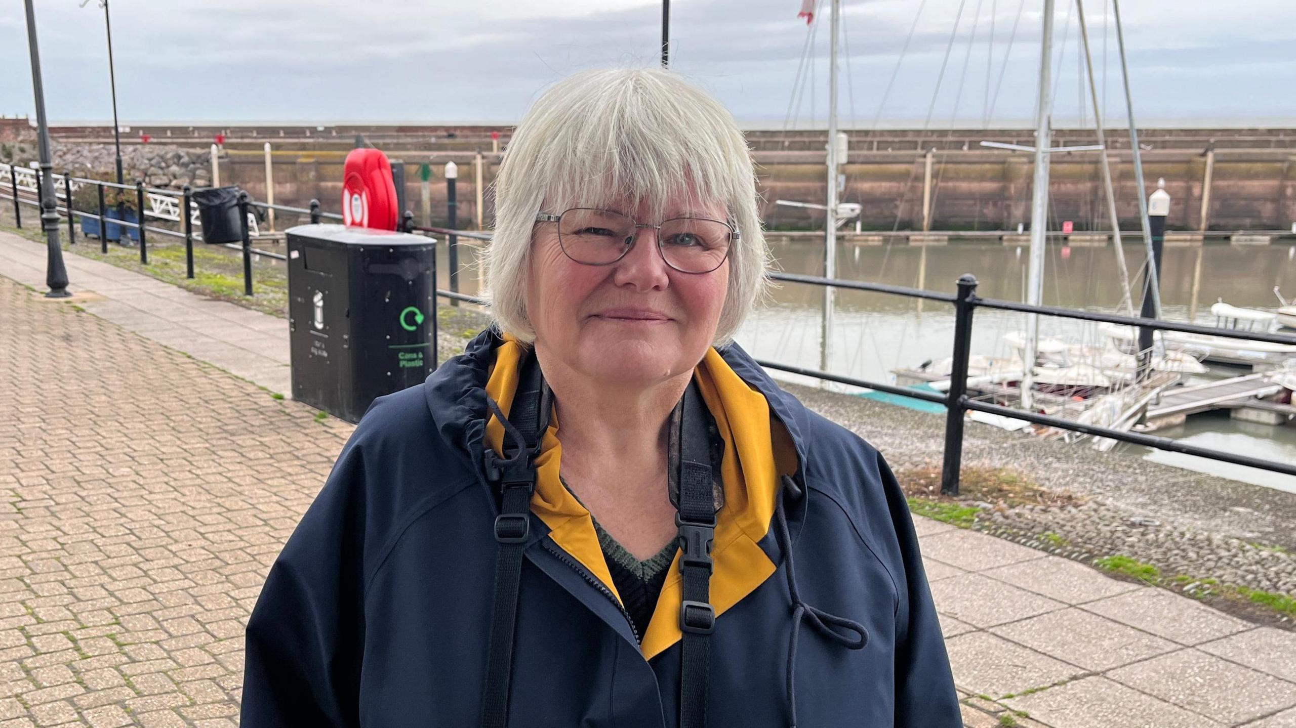 Liz Elmont is standing on a boardwalk by the harbour in Watchet. She is wearing a blue raincoat and is smiling at the camera.
