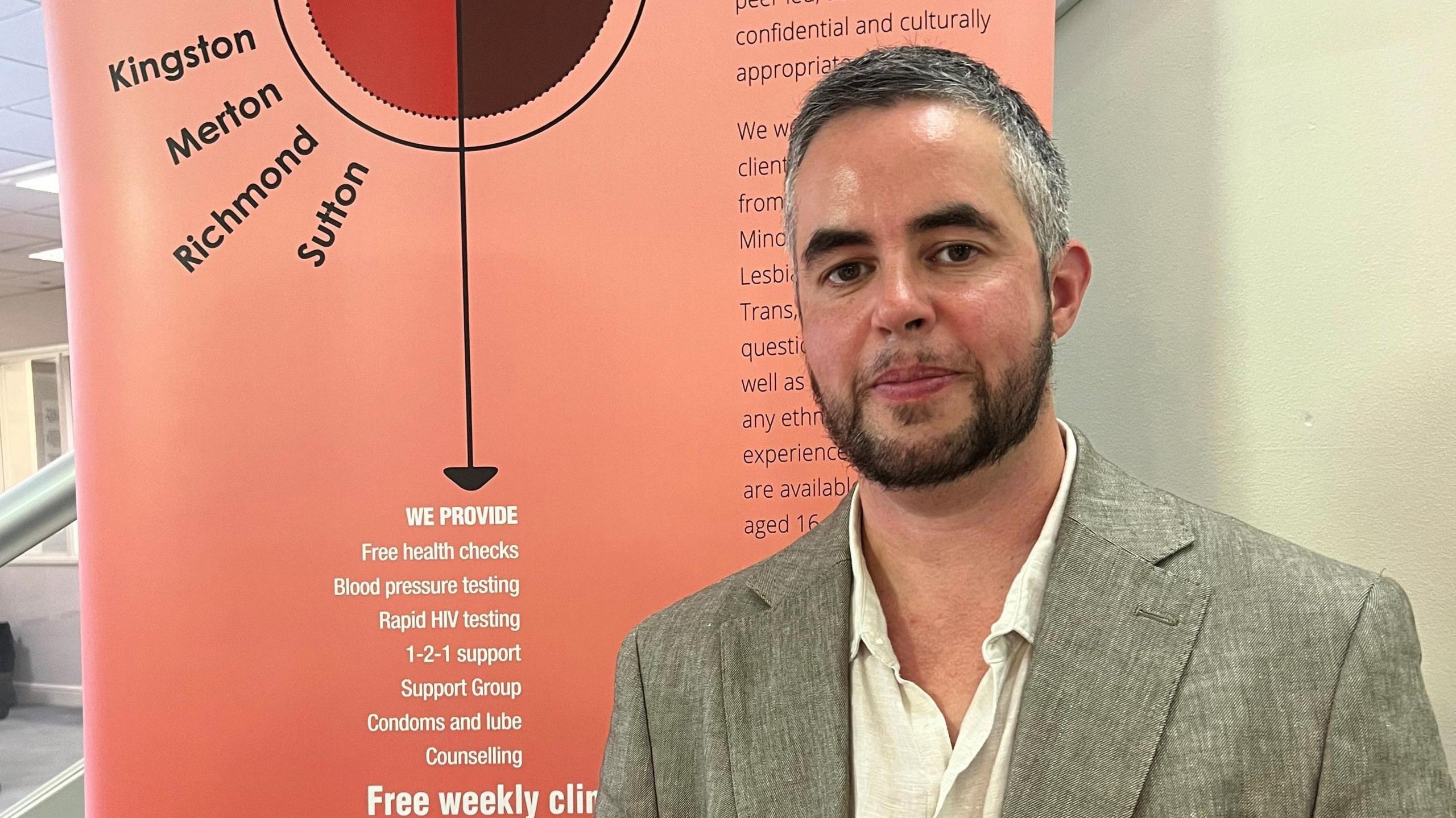 Joel Robinson, who has greying hair, a beard and dark eyebrows, is standing in front of a sign that provides information about the charity's outreach clinics in London. He is wearing a light-coloured shirt and a grey suit jacket and is looking at the camera.