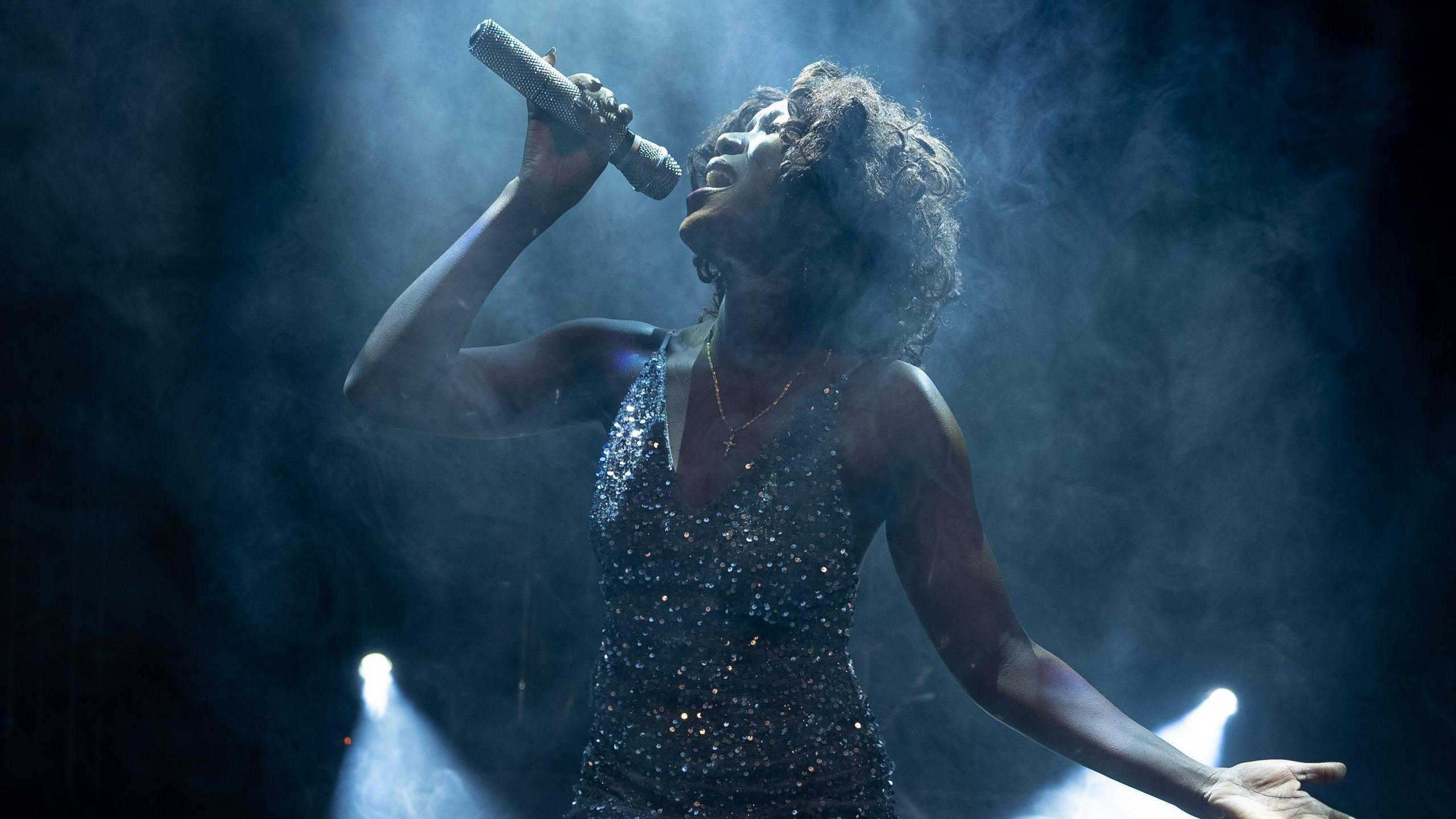 A woman playing Whitney Houston in a show called Queen of the Night at the Bristol Hippodrome. She is wearing a sparkling dress and is holding a microphone above her head as she sings