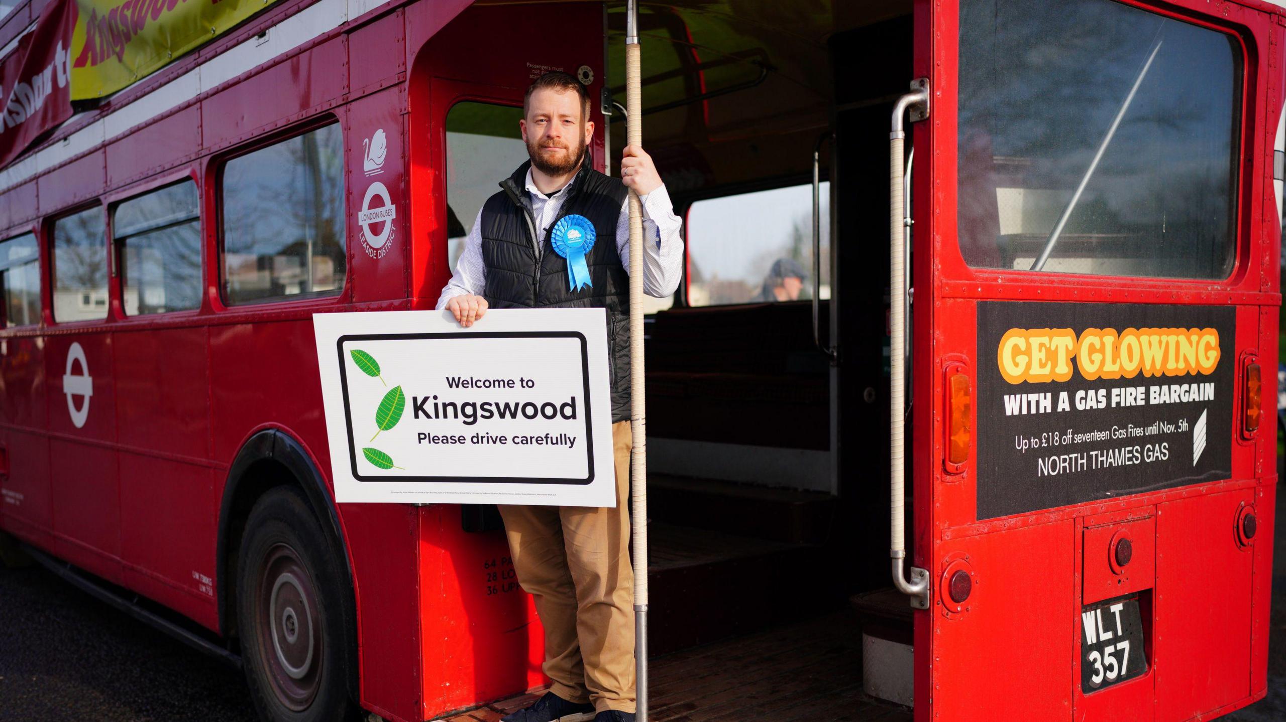 Councillor Sam Bromiley on the back of a bus holding a sign that says "welcome to Kingswood"